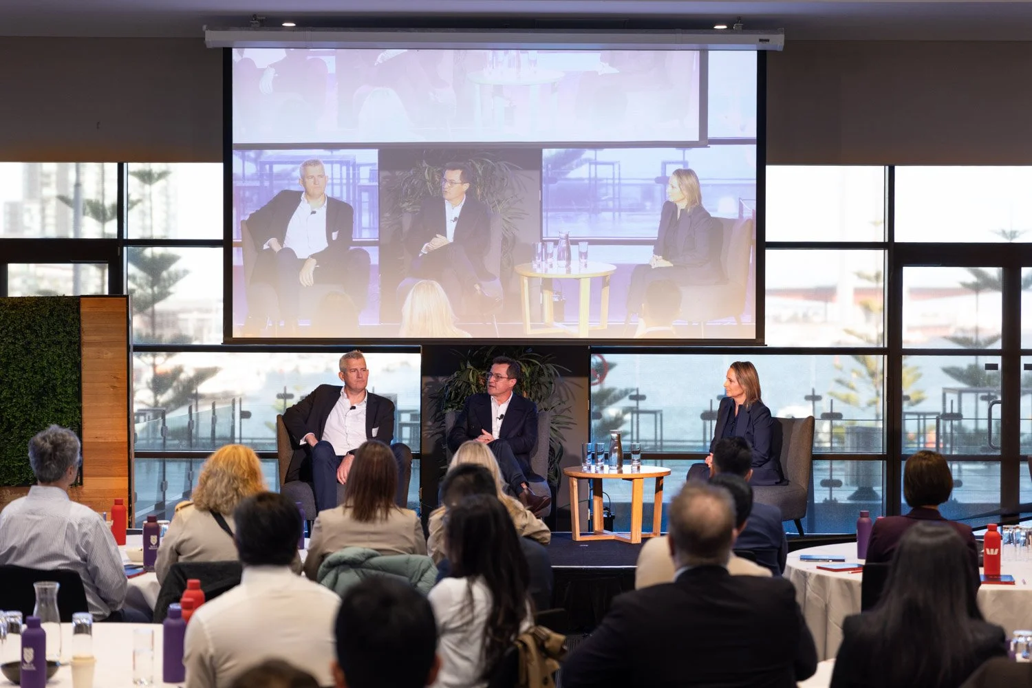 Panel discussion at conference, with three speakers on stage and audience watching inside a room with large windows overlooking a marina.