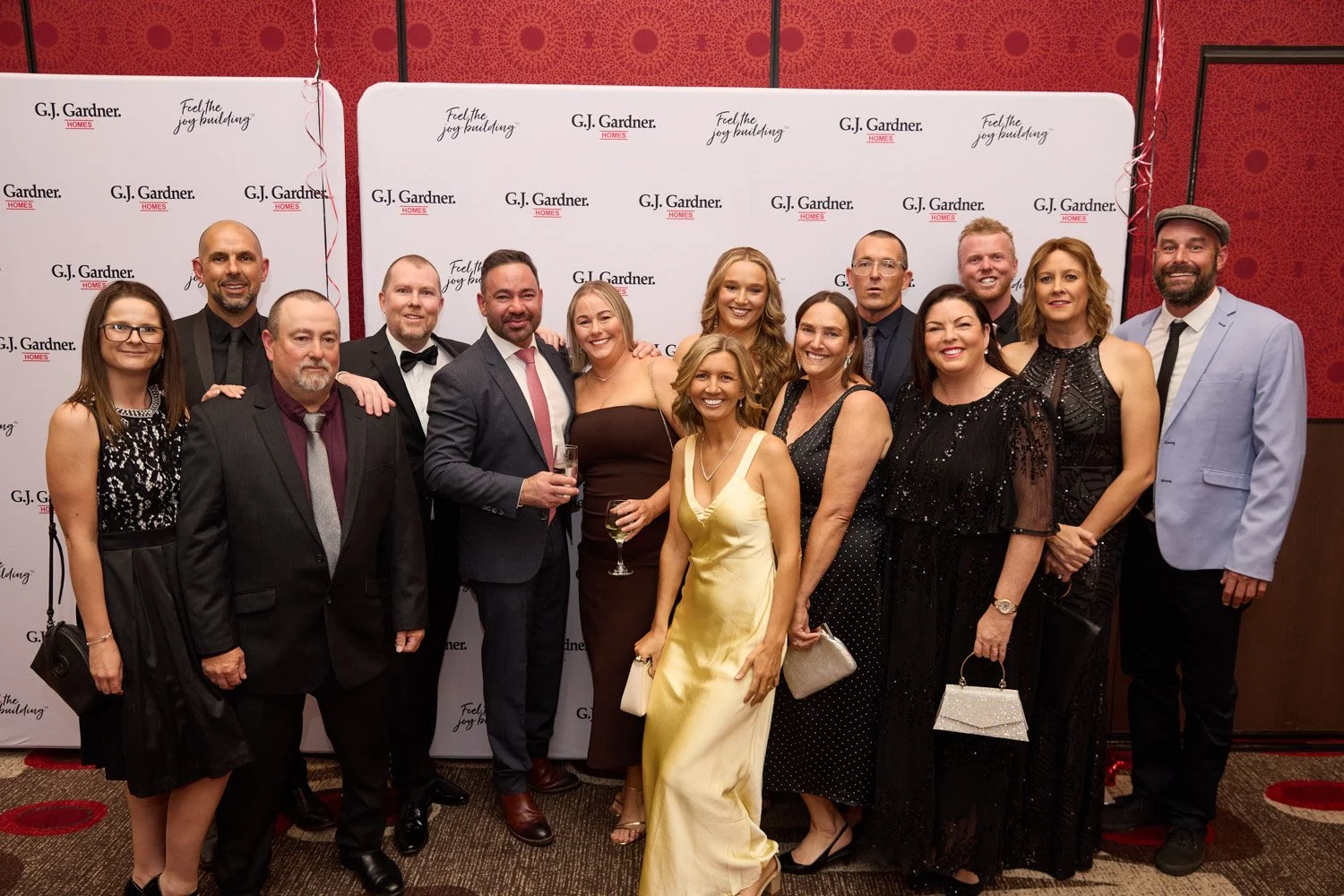 A group of 14 people dressed in formal attire posing together at a G.J. Gardner Homes event, with a white backdrop featuring the company logo and slogan, in an indoor setting with red patterned walls.