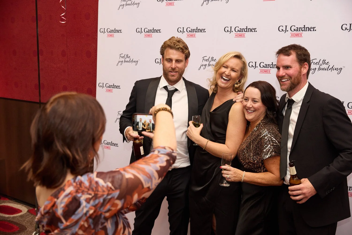 Four adults in formal attire smiling and posing for a photo at an event, with a woman taking their picture with a smartphone. They are holding drinks and standing in front of a branded backdrop.