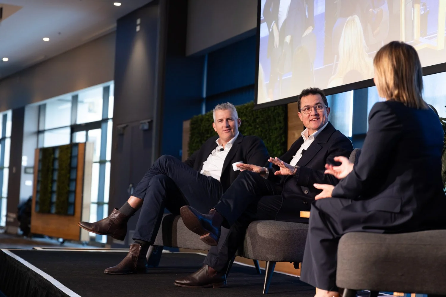 Three people wearing business suits sitting on stage in front of a large screen, engaging in a panel discussion with a woman. The stage is indoors with large windows in the background.