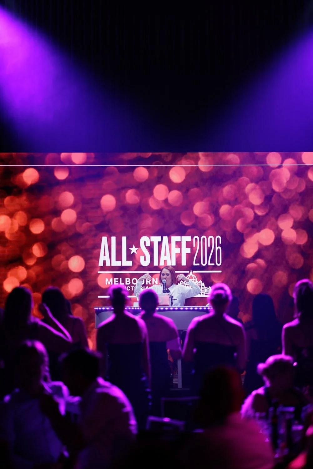 A woman at a podium speaking to an audience at the All Staff 2026 event in Melbourne, with a large digital screen behind displaying the event name and festive background.