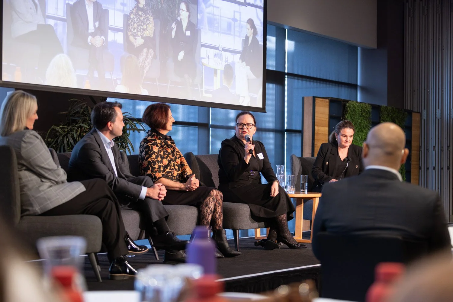 Panel discussion at a conference with five women and one man on stage, one woman speaking into a microphone, large screen behind displaying a live feed of the panel, in a modern room with windows and green wall decor.