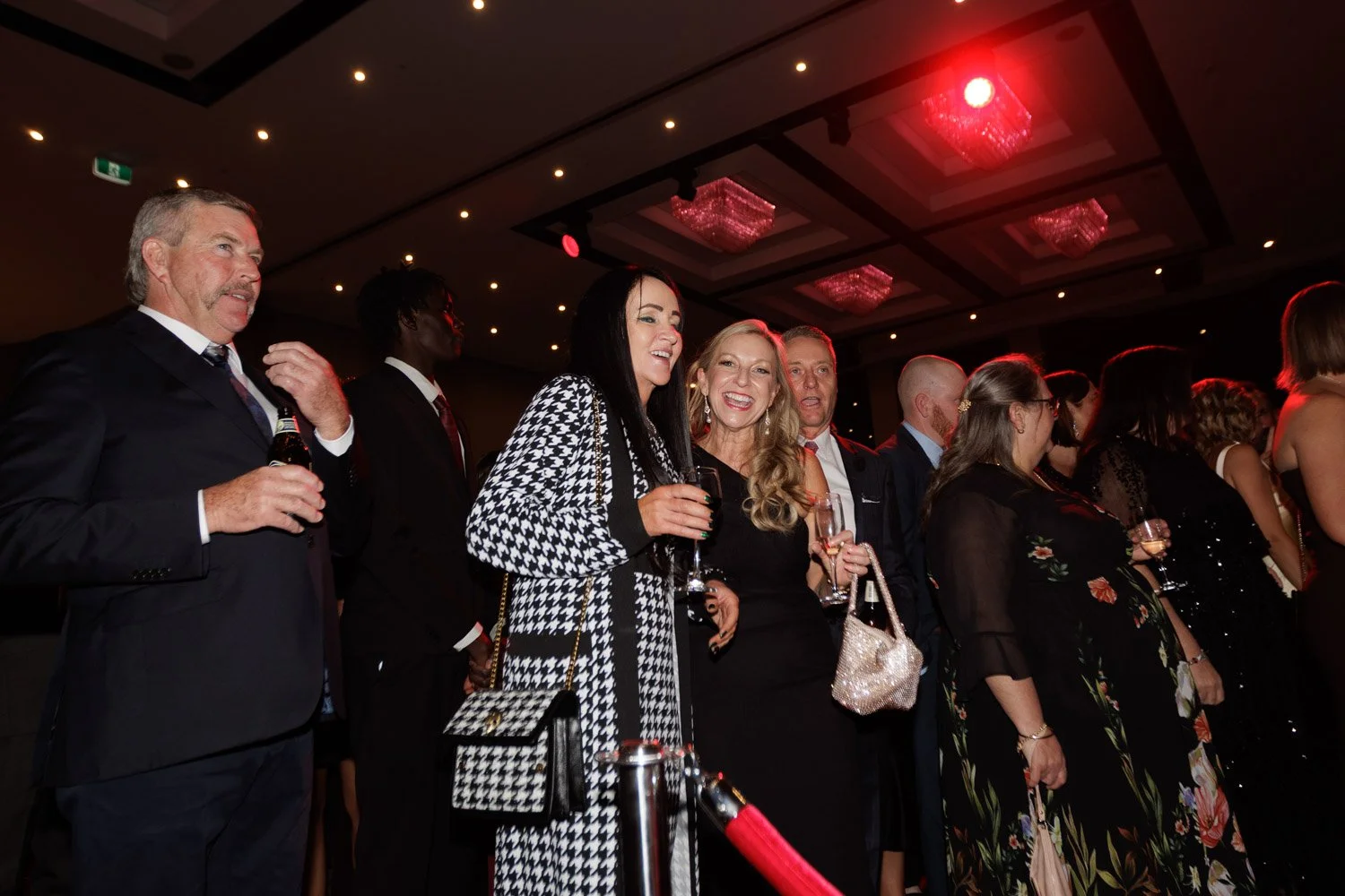 Group of people dressed in formal attire at a party or event, standing in a line with some holding drinks, in a dimly lit room with ceiling chandeliers and red lighting.
