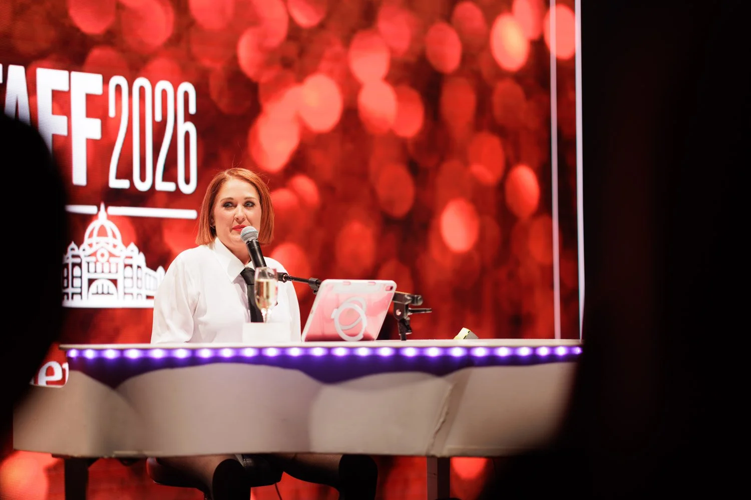 A woman with red hair wearing a white shirt and black tie is speaking into a microphone at a podium with a drink, in front of a red bokeh background displaying the text 'AFE 2020' and a white building logo.
