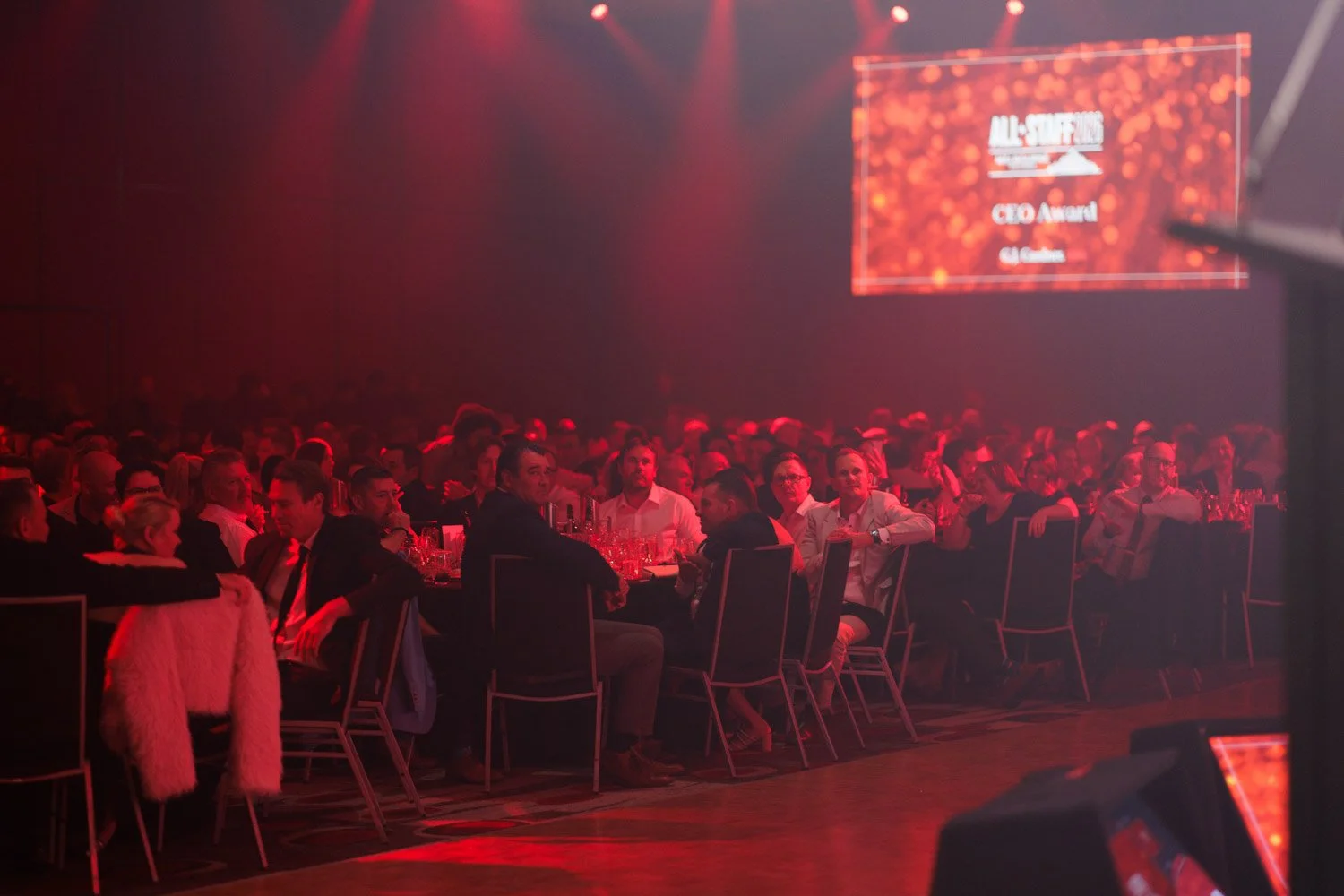 An audience sitting at multiple round tables during an award ceremony, with a large screen displaying 'All-Stars' and 'CEO Award' in the background, under red lighting.