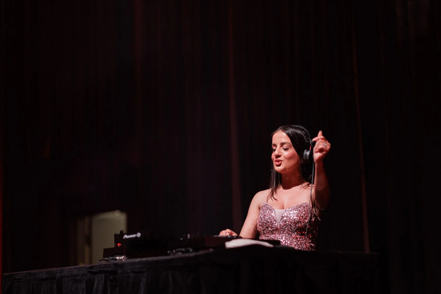 A woman wearing a glittery pink top is DJing with headphones on, standing behind a DJ console, with dark curtains in the background, seemingly enjoying music.