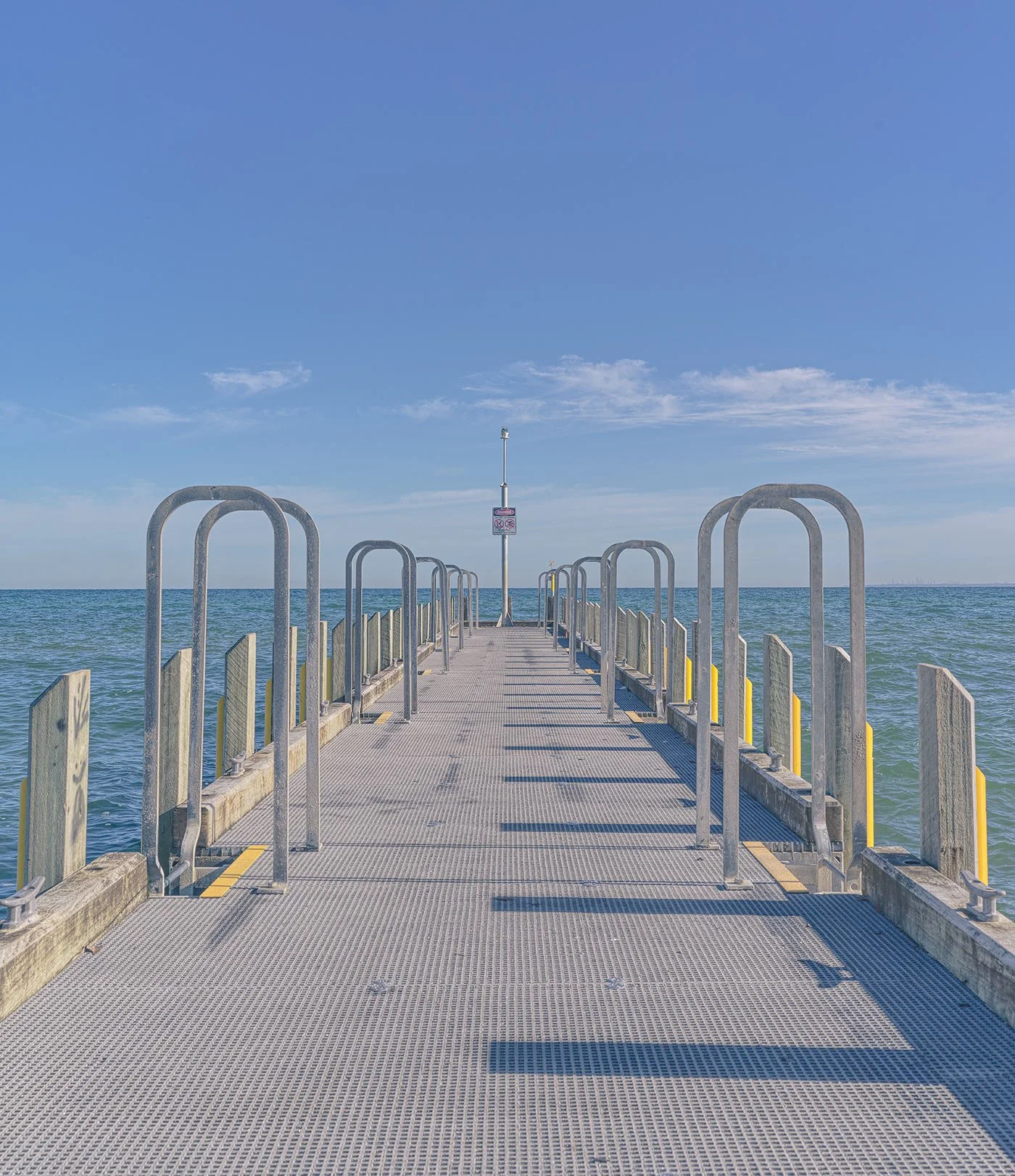 A metal dock extending into the ocean with handrails on both sides and a warning sign, under a blue sky with a few clouds.