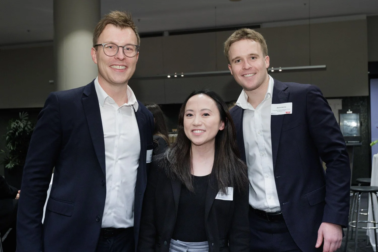 Three people, two men and one woman, smiling and posing together at an indoor event, all wearing business attire including white shirts and dark blazers.