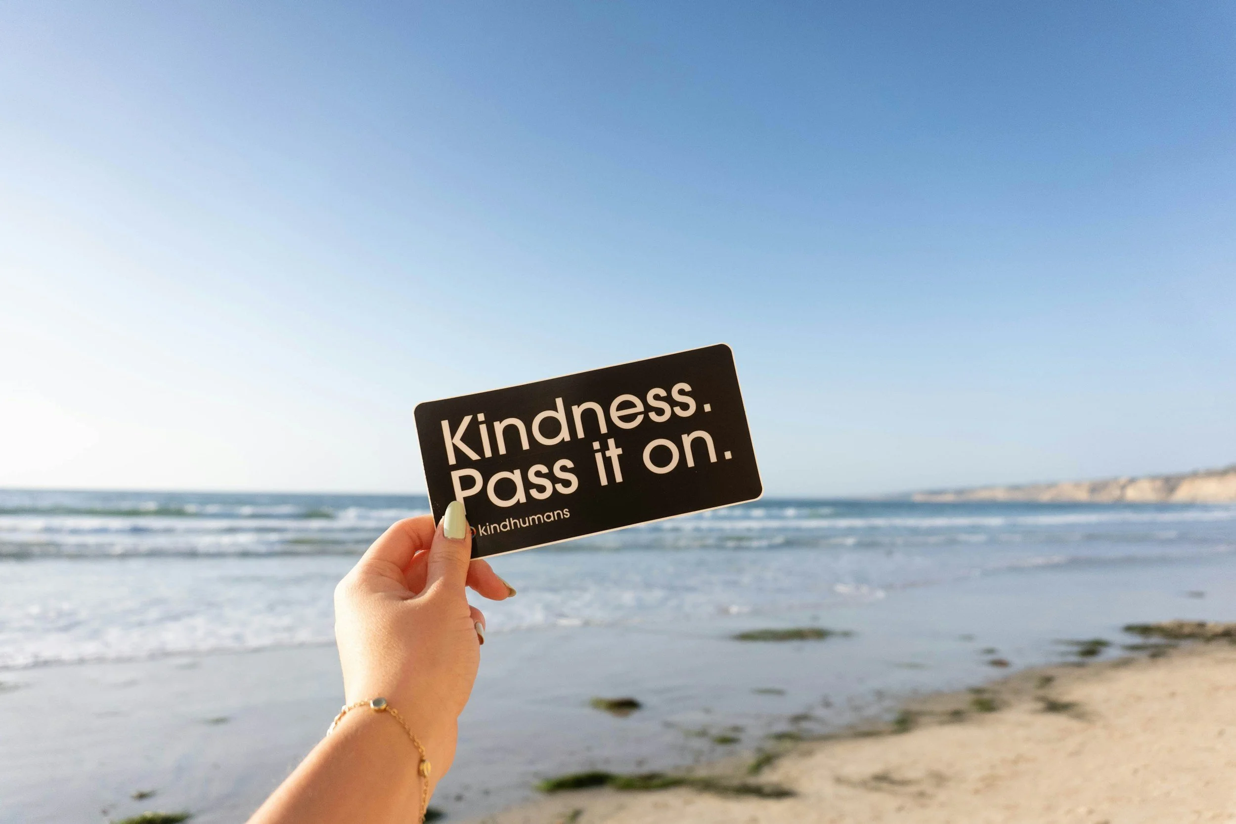 kindness. pass it on. is written on a post card and held up by the beach. the ocean waves are glistening in the sunlight behind. this creates a backdrop for the upcoming blog about why sharing kind words may matter more than we think.