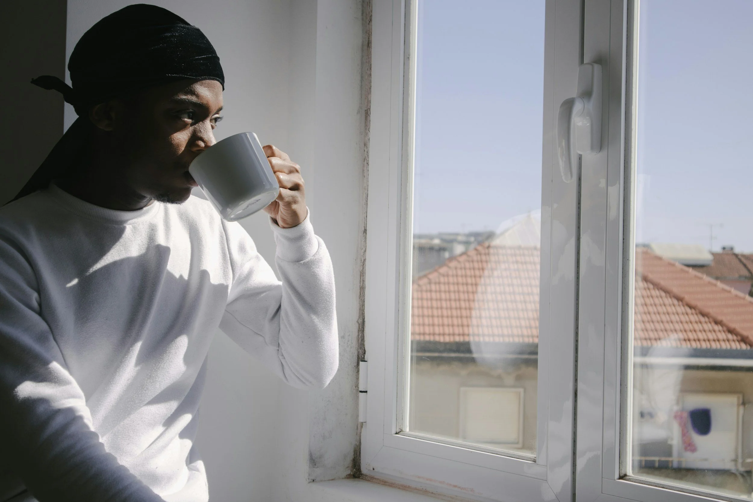 a man with his coffee cup to his mouth stares out of his sun soaked window