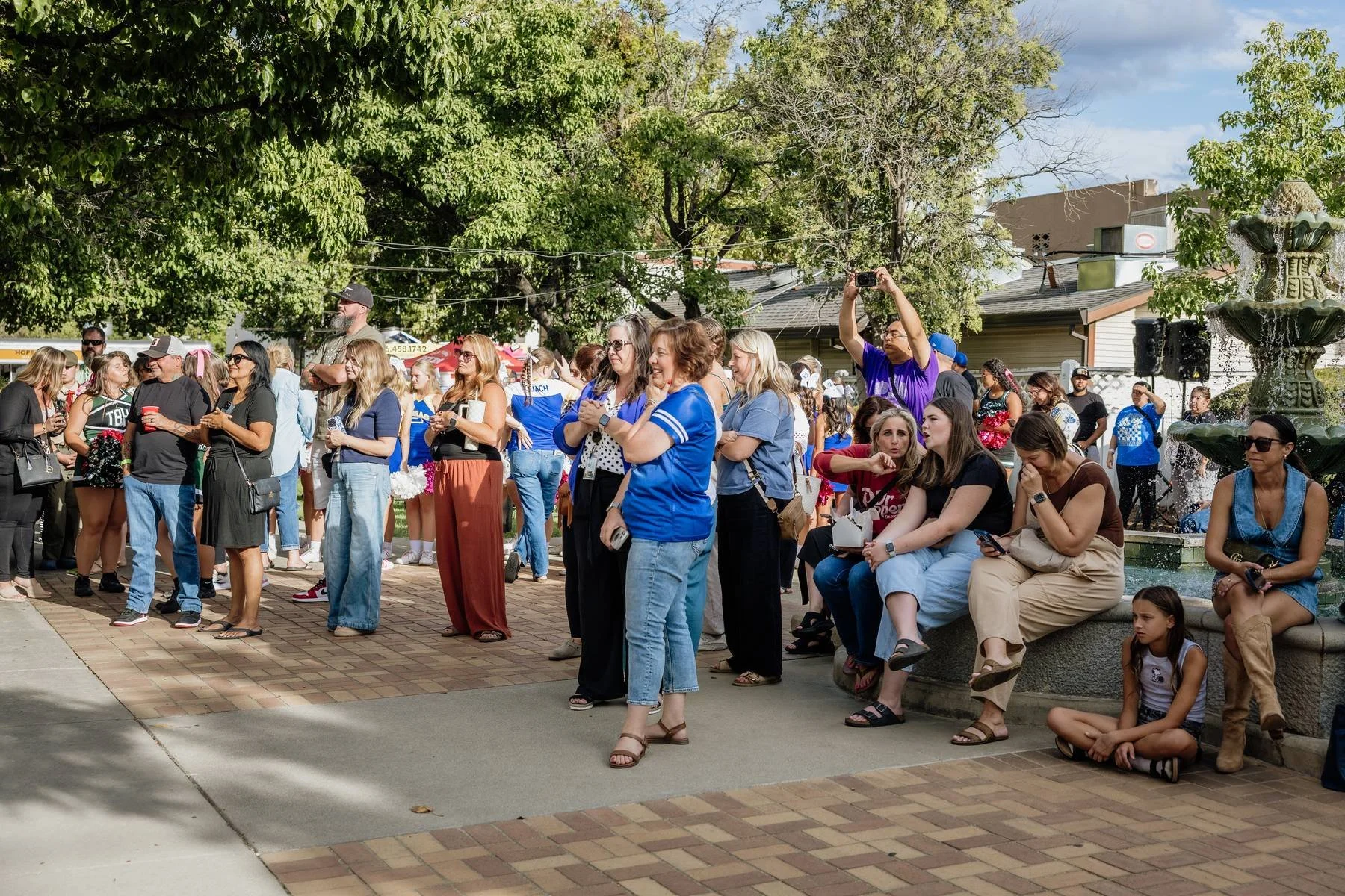 People gathered outdoors in a park-like setting, some sitting on a fountain and others standing, with trees and buildings in the background.