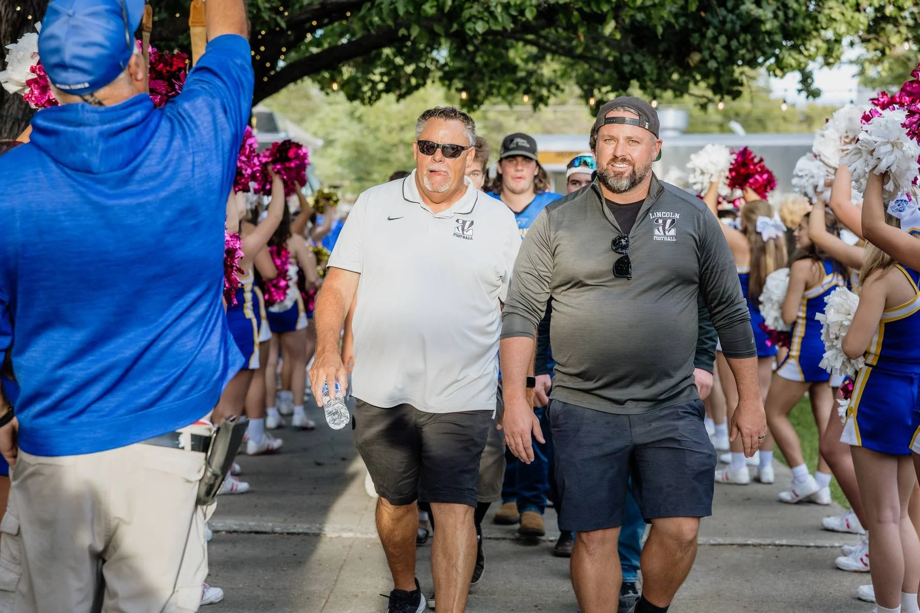 Two men walking through a cheerleading squad arranged in a tunnel, with cheerleaders holding pom-poms on either side. One man is wearing a white polo shirt, sunglasses, and shorts, while the other is in a black athletic shirt and cap.