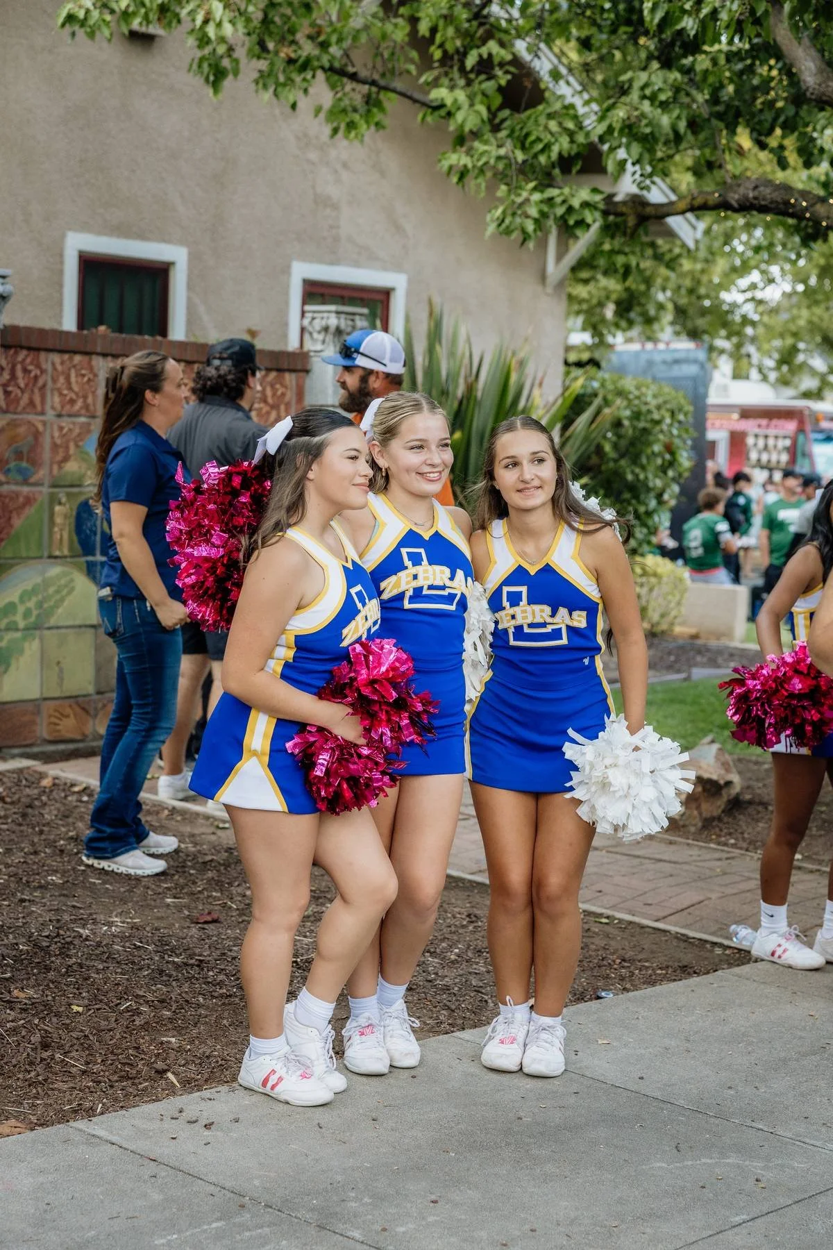 Three female cheerleaders in blue and yellow uniforms with pom-poms, standing outdoors in front of a beige building and tree, smiling and posing for a photo.