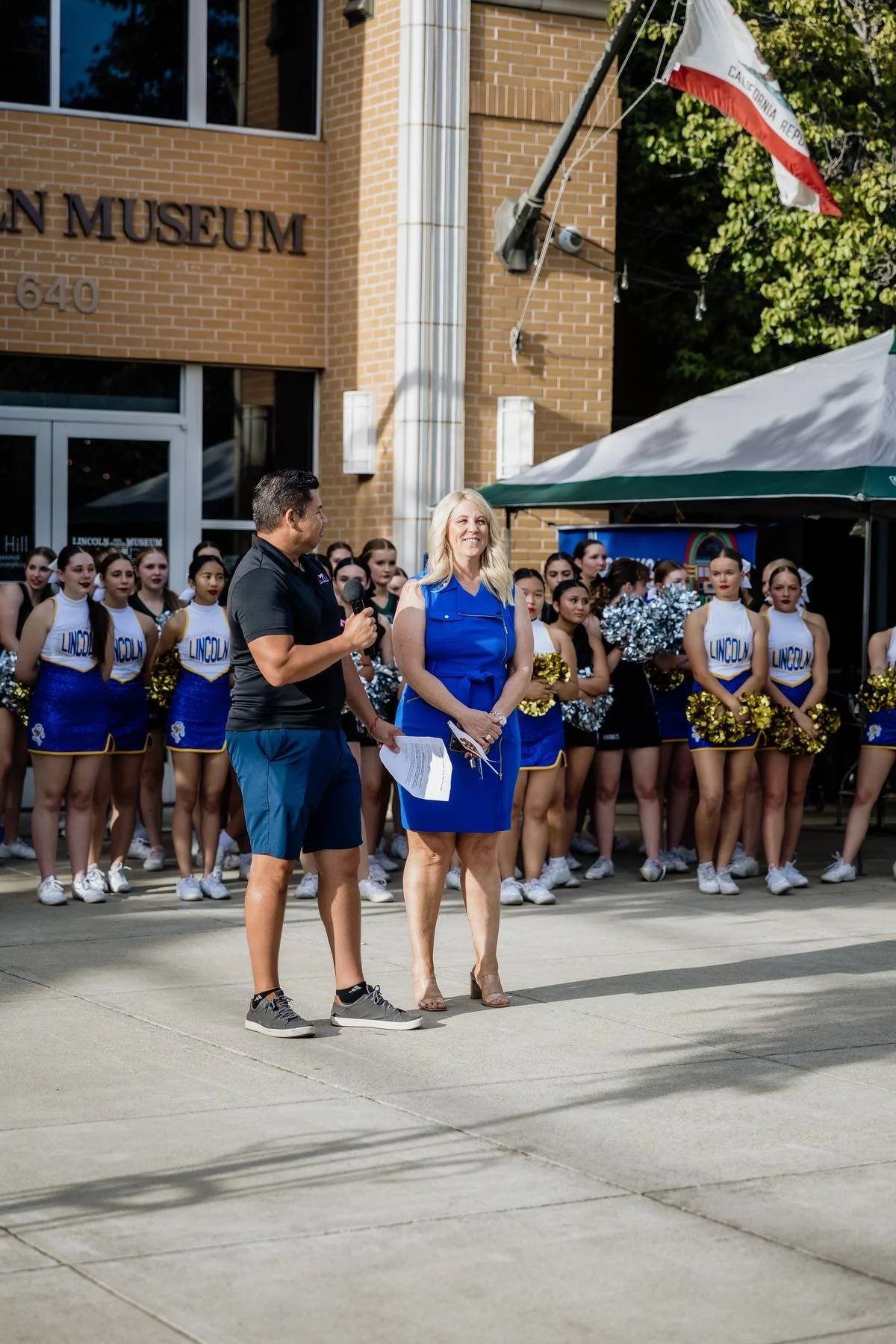 A group of cheerleaders in blue and white uniforms standing outside Lincoln Museum. Two individuals are speaking in front of the cheerleaders, one holding a microphone and the other holding papers, at an outdoor event.