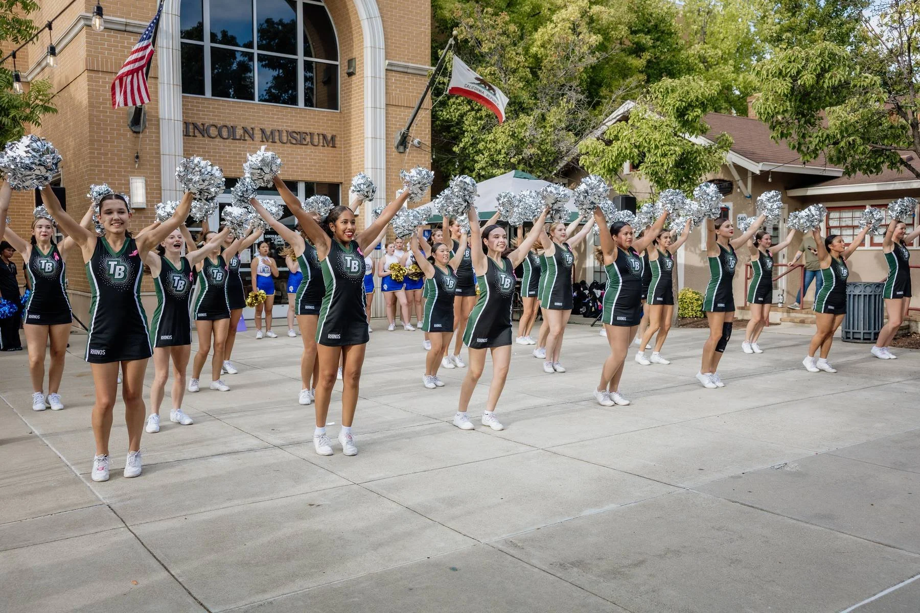 Cheerleaders performing a routine with silver pom-poms in front of the Lincoln Museum, with a group of other cheerleaders in white and blue uniforms in the background.