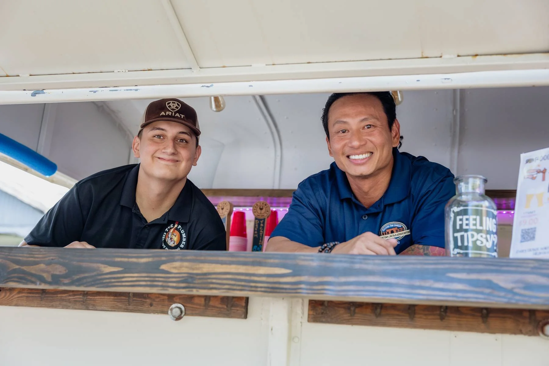 Two smiling men leaning over a counter at a food stand, one wearing a black shirt and brown cap, the other in a blue shirt, with colorful ribbons and a sign that reads "Feeling Tipsy?" in the background.