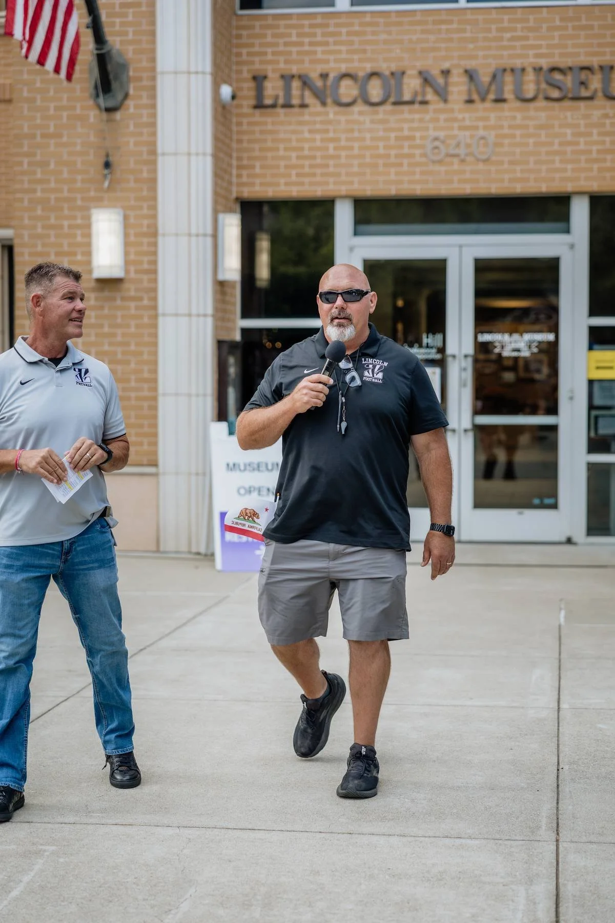 Two men, one in a gray shirt and the other in a black shirt and shorts, standing in front of the Lincoln Museum; the man in black holding a microphone, the other holding papers, with the museum entrance behind them.