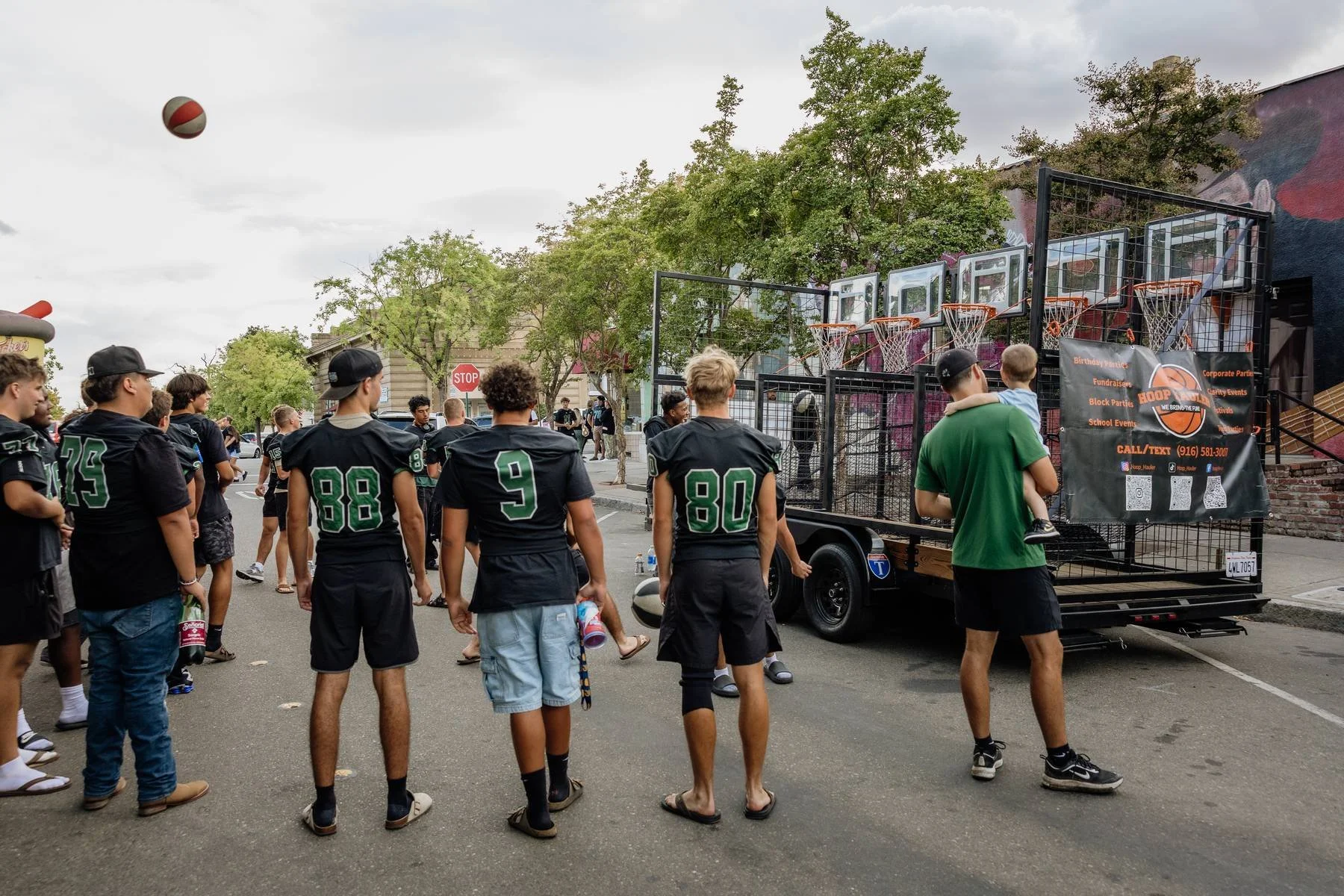A group of young men in football jerseys gathered around a mobile basketball court on a city street, with some watching and one man holding a child, in a scene with trees and buildings in the background.