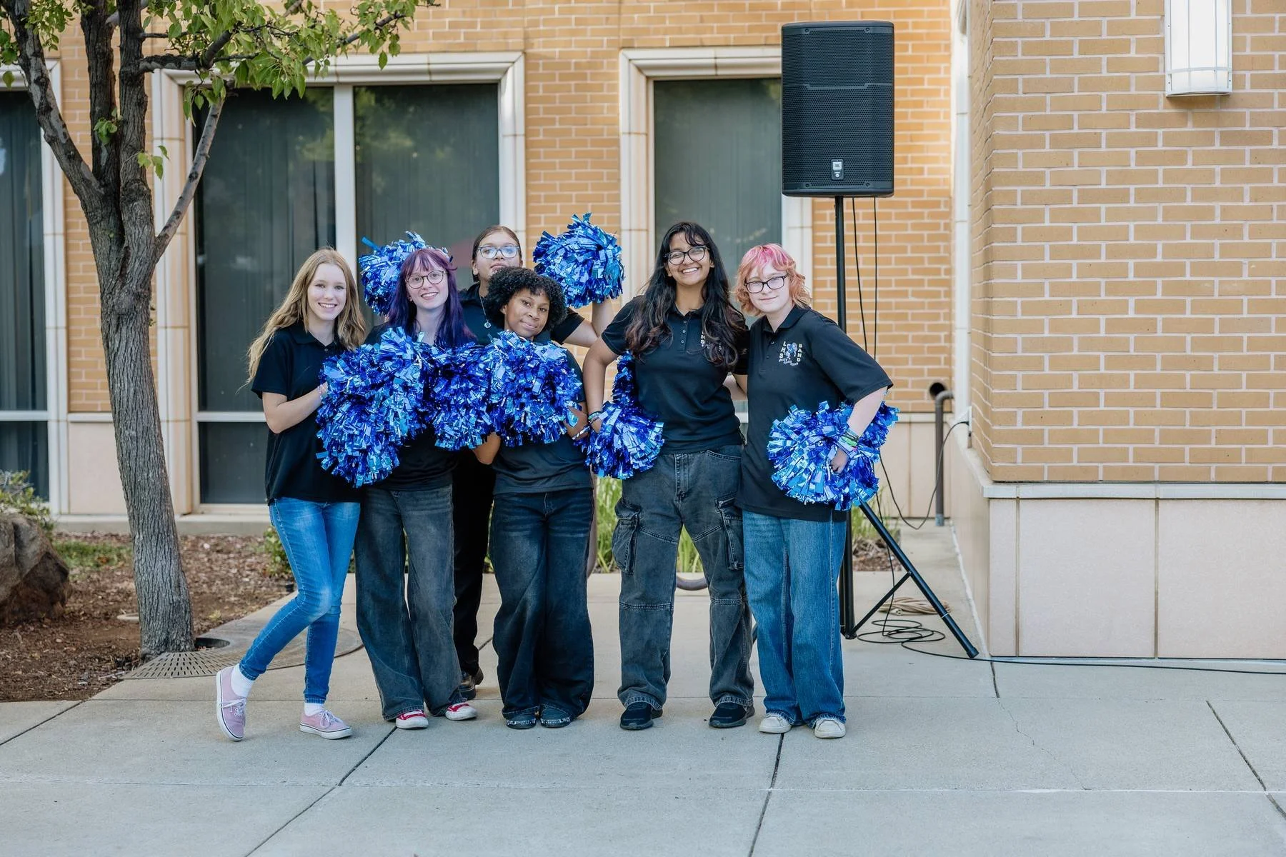 Group of six girls standing outdoors, holding blue pom-poms, smiling, in front of a brick building with a speaker and trees.