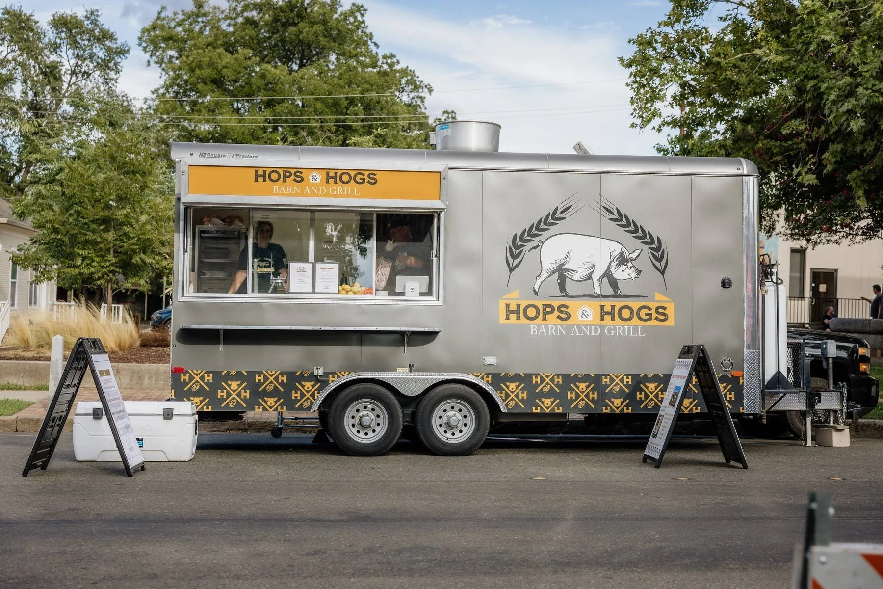 Food truck named 'Hops & Hogs Barn and Grill' with a window serving customers, featuring a pig logo with wheat stalks, parked on the street with signs and a white cooler nearby.