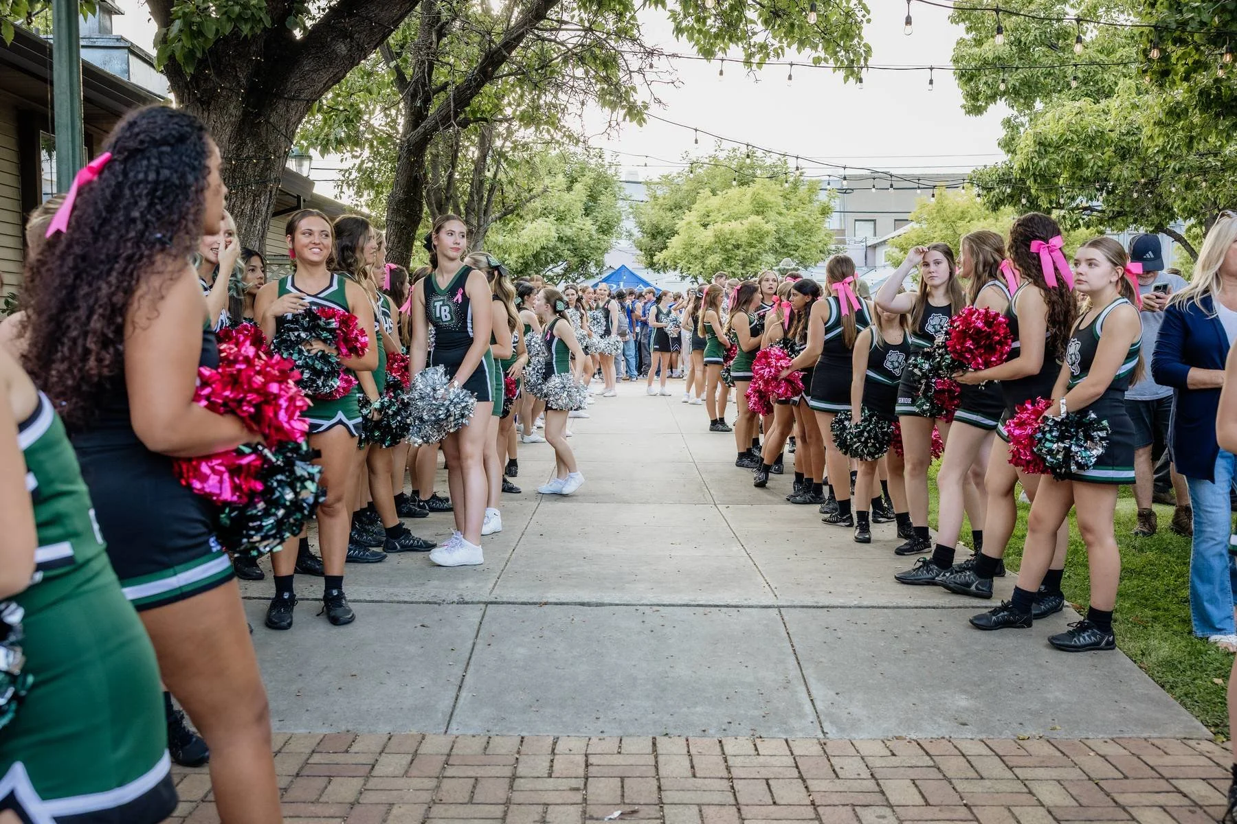 A group of cheerleaders in black and green uniforms are standing in two lines facing each other on a sidewalk, holding pink and silver pom-poms, with some pink ribbons in their hair. There are spectators and trees in the background.