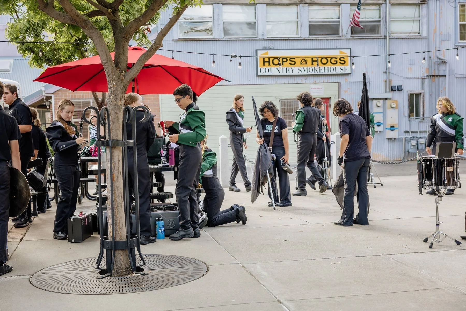 School band students preparing and setting up instruments outside a country smokehouse with a sign reading 'Hops & Hogs'.