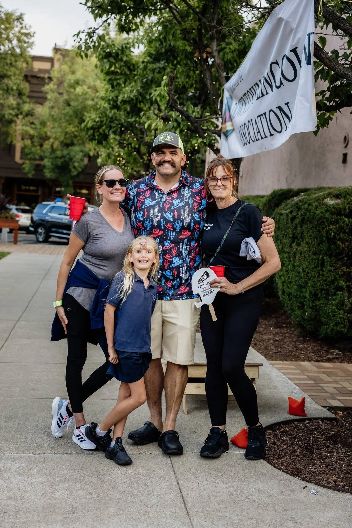 Group of four people and a child at an outdoor event, standing on a sidewalk with trees and a building in the background. They are smiling, holding drinks and a small sign, with a flag partially visible behind them.