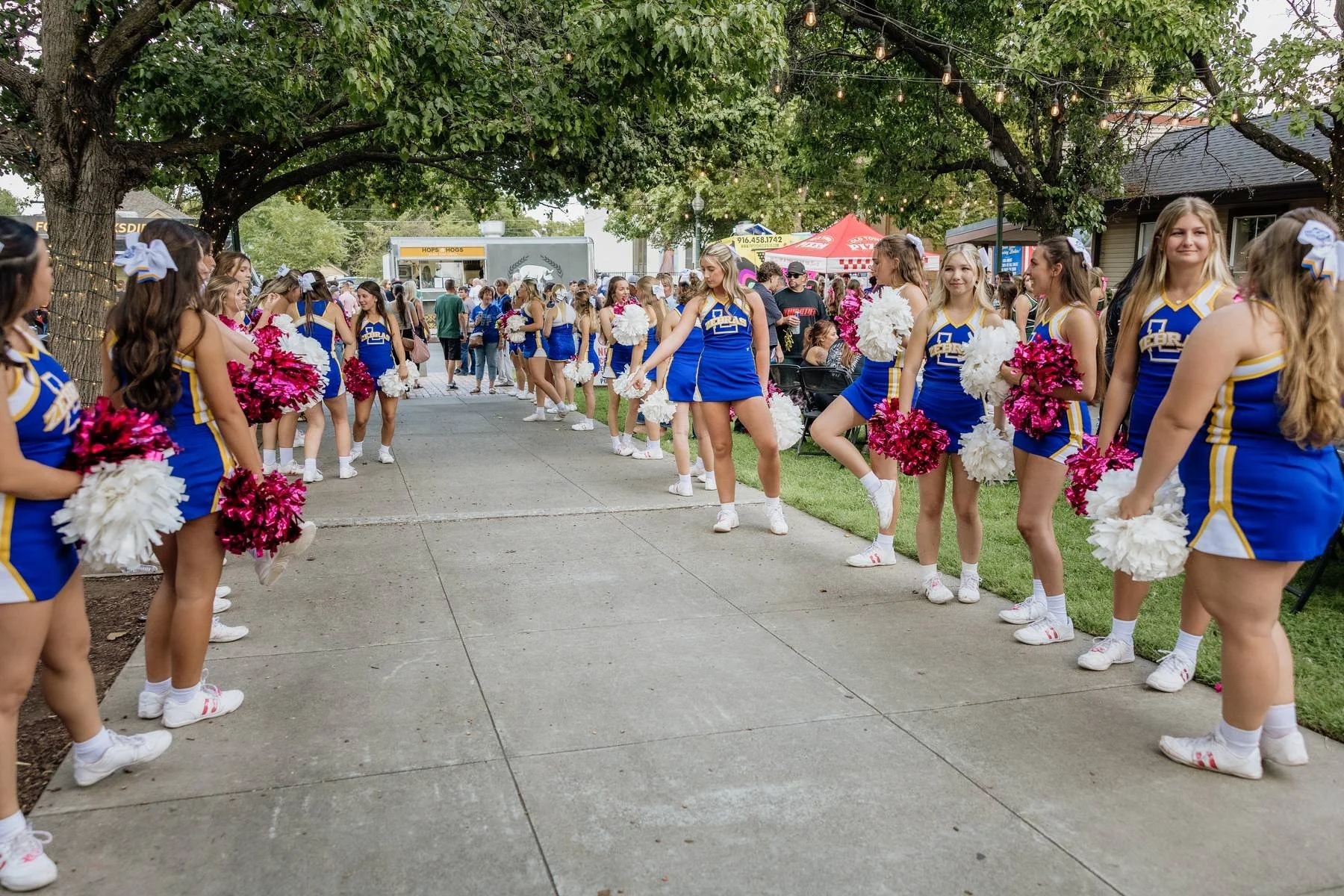 Line of cheerleaders in blue uniforms holding pink and white pom-poms at a street fair or outdoor event with trees and food stalls in the background.