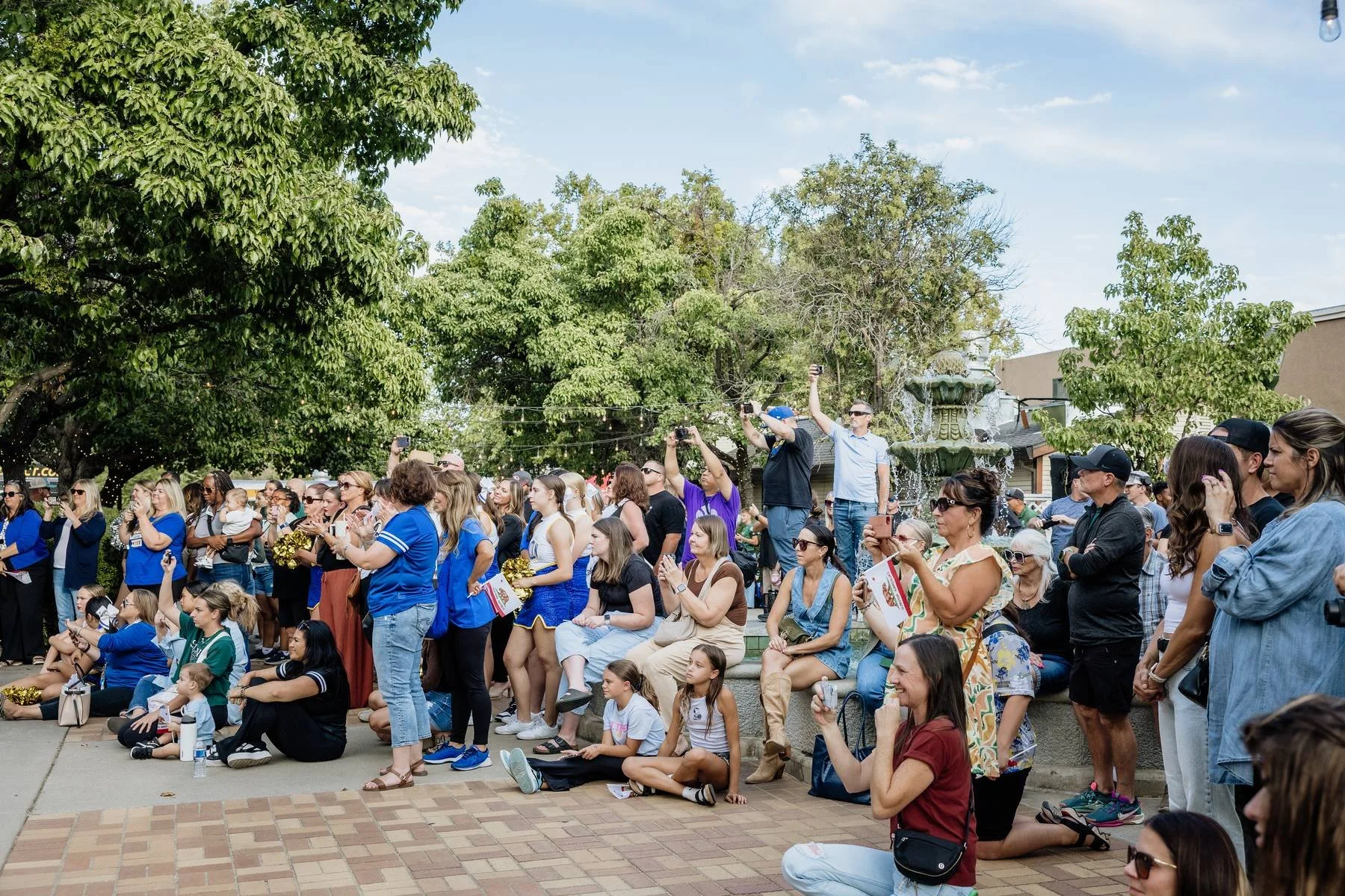 Crowd of people gathered outdoors, some taking photos, others sitting or standing, with a fountain and trees in the background.