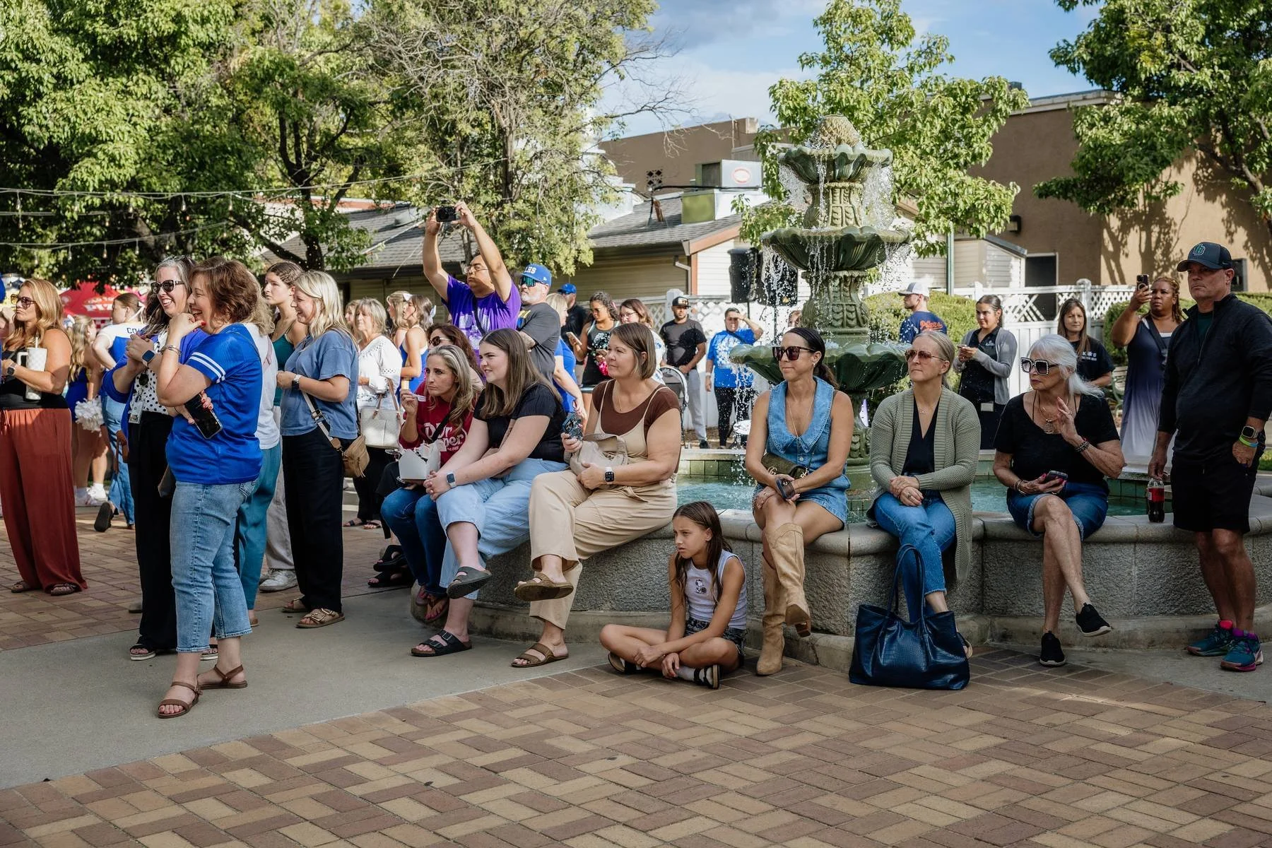 Crowd of people gathered in front of a fountain in an outdoor setting with trees and buildings in the background. Some people are seated, some standing, many holding phones or drinks, and a few taking photos or videos.
