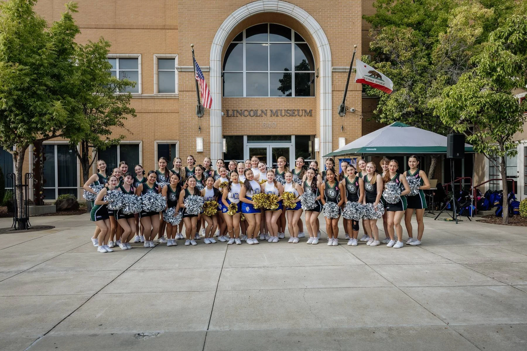 Group of cheerleaders posing in front of the Lincoln Museum, holding pom-poms, with some wearing blue and white uniforms and others in black and teal uniforms.