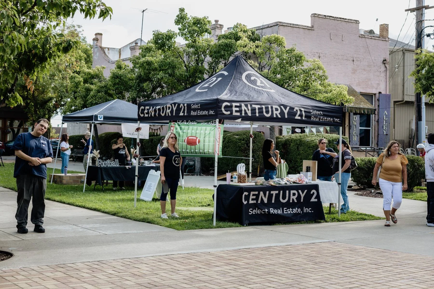 People gathered around real estate tents on a sidewalk in a park, with trees and buildings in the background, some browsing items and others socializing.