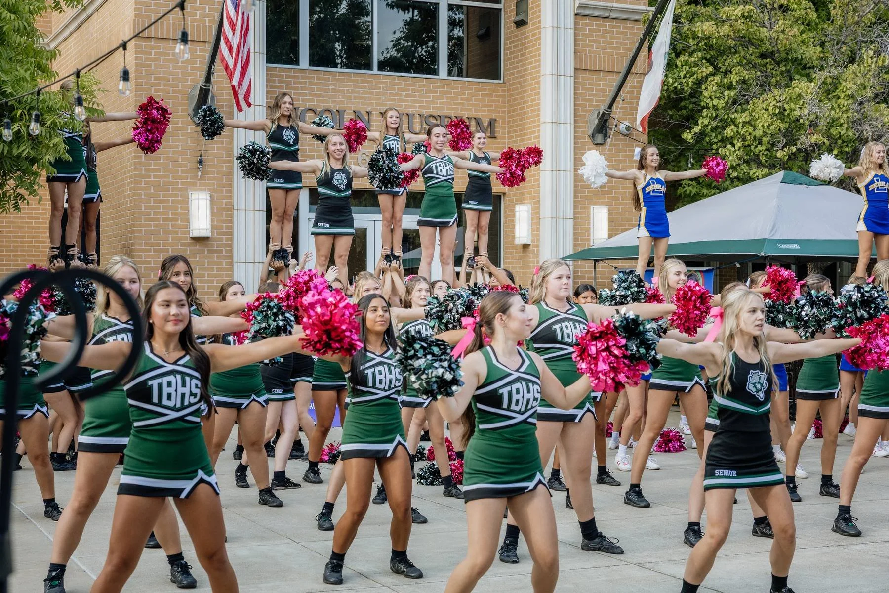School cheerleaders performing a cheer routine outdoors in front of a brick school building with an American flag and a school flag. They are wearing green and black uniforms with