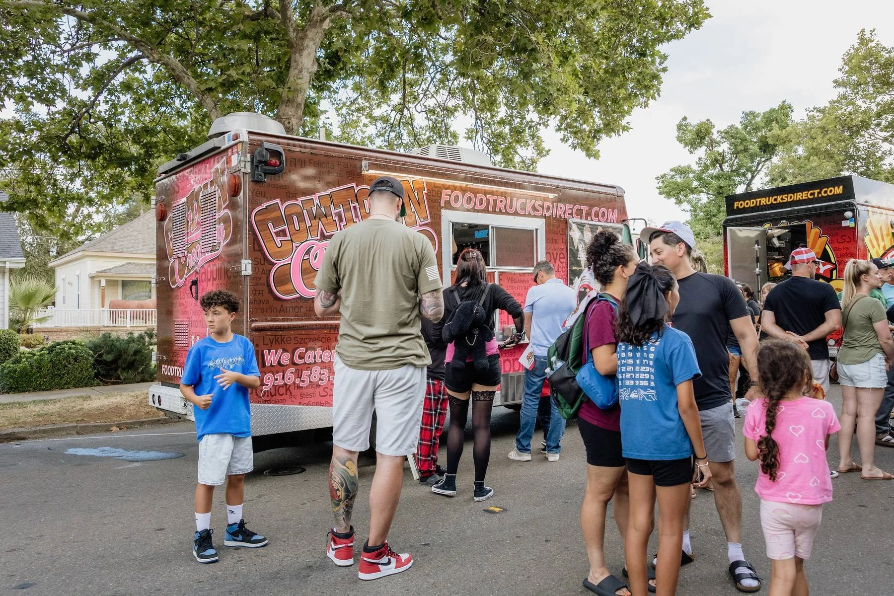 People standing in line at two food trucks parked on a street, surrounded by trees and residential houses, on a partly cloudy day.