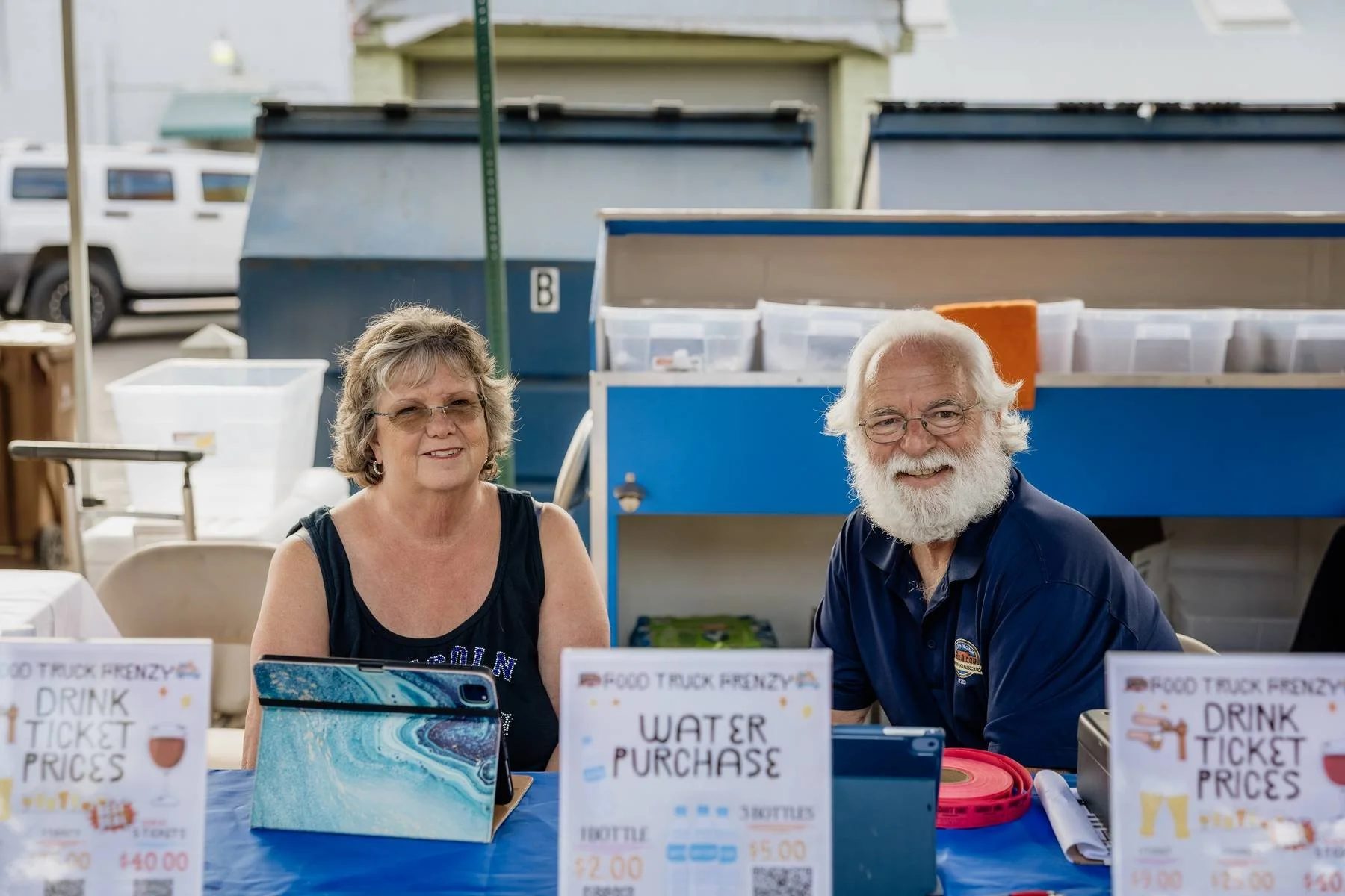 A smiling elderly man with glasses and a white beard and a woman with glasses sitting at a table outdoors at a food truck event.
