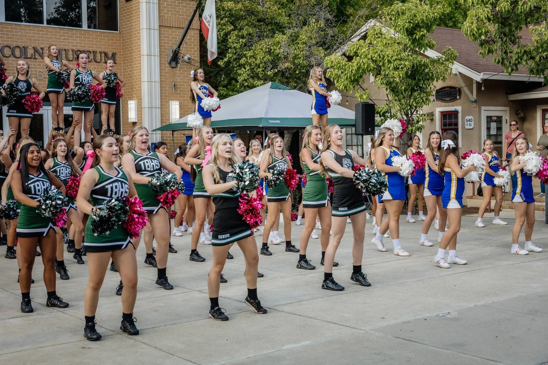 A group of cheerleaders performing a routine outdoors on a pavement in front of a building with a sign that reads 'COLLINSUM.' The cheerleaders are wearing uniforms in green, black, and dark gray, and blue and yellow with some holding pink, white, an