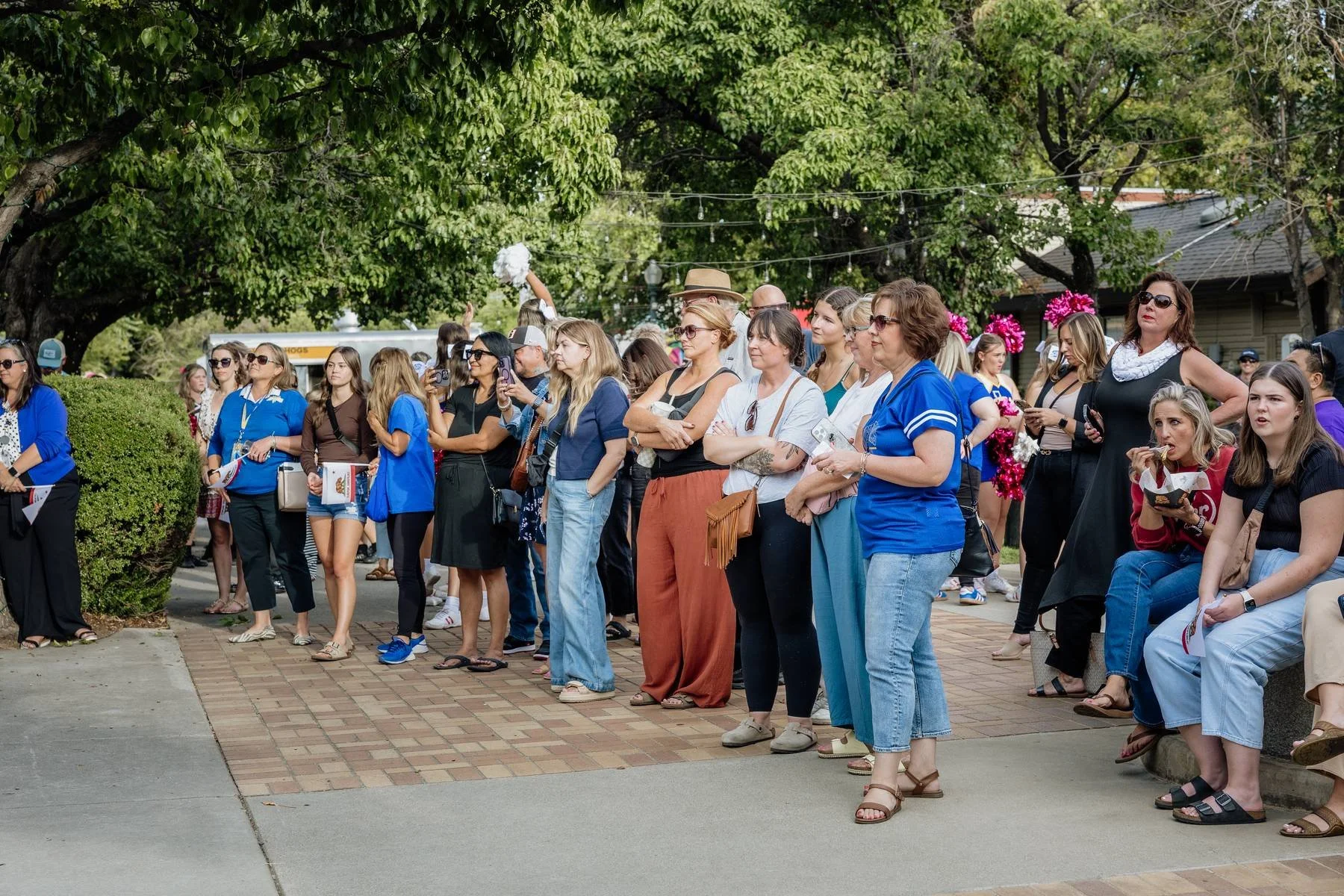 A crowd of people, mostly women, gathered outdoors on a sidewalk, some watching an event and others taking photos, with trees and string lights overhead.
