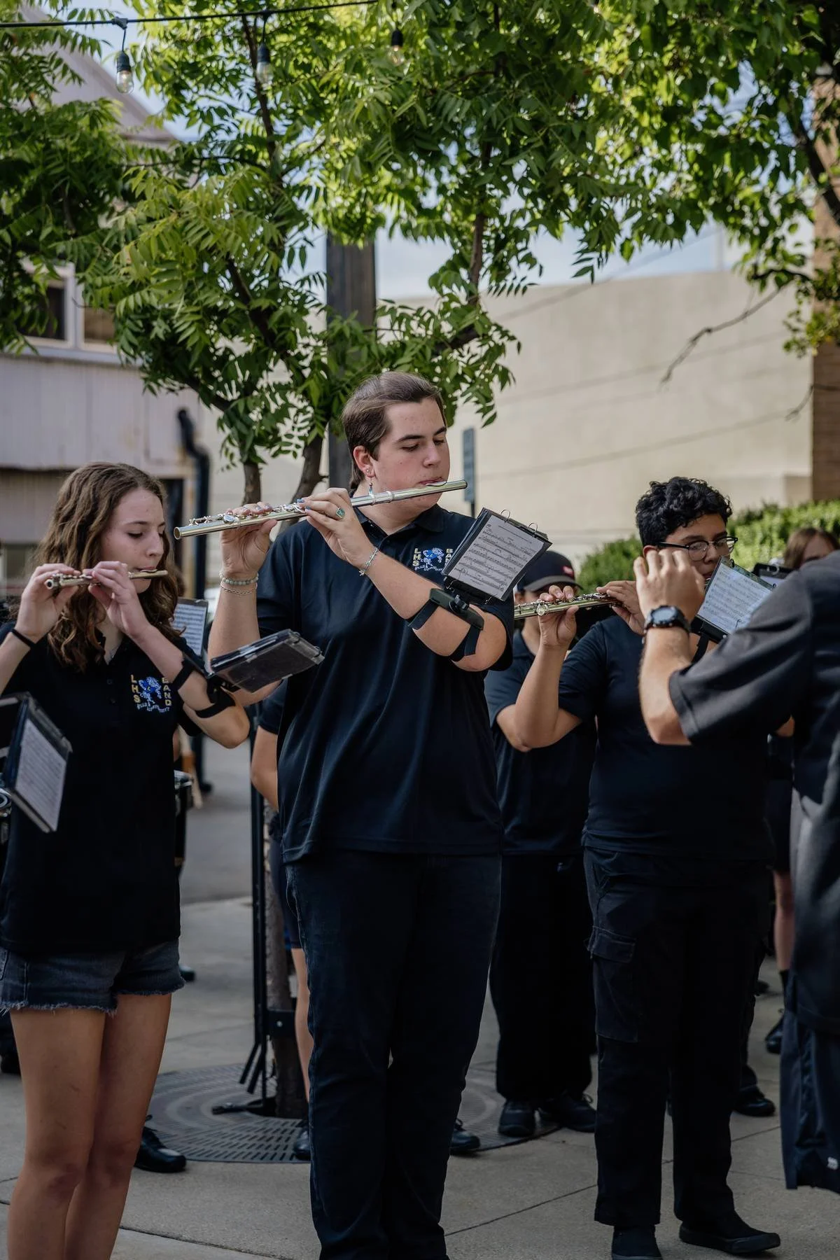 Group of young people playing flutes outdoors during the day, under a tree with string lights.