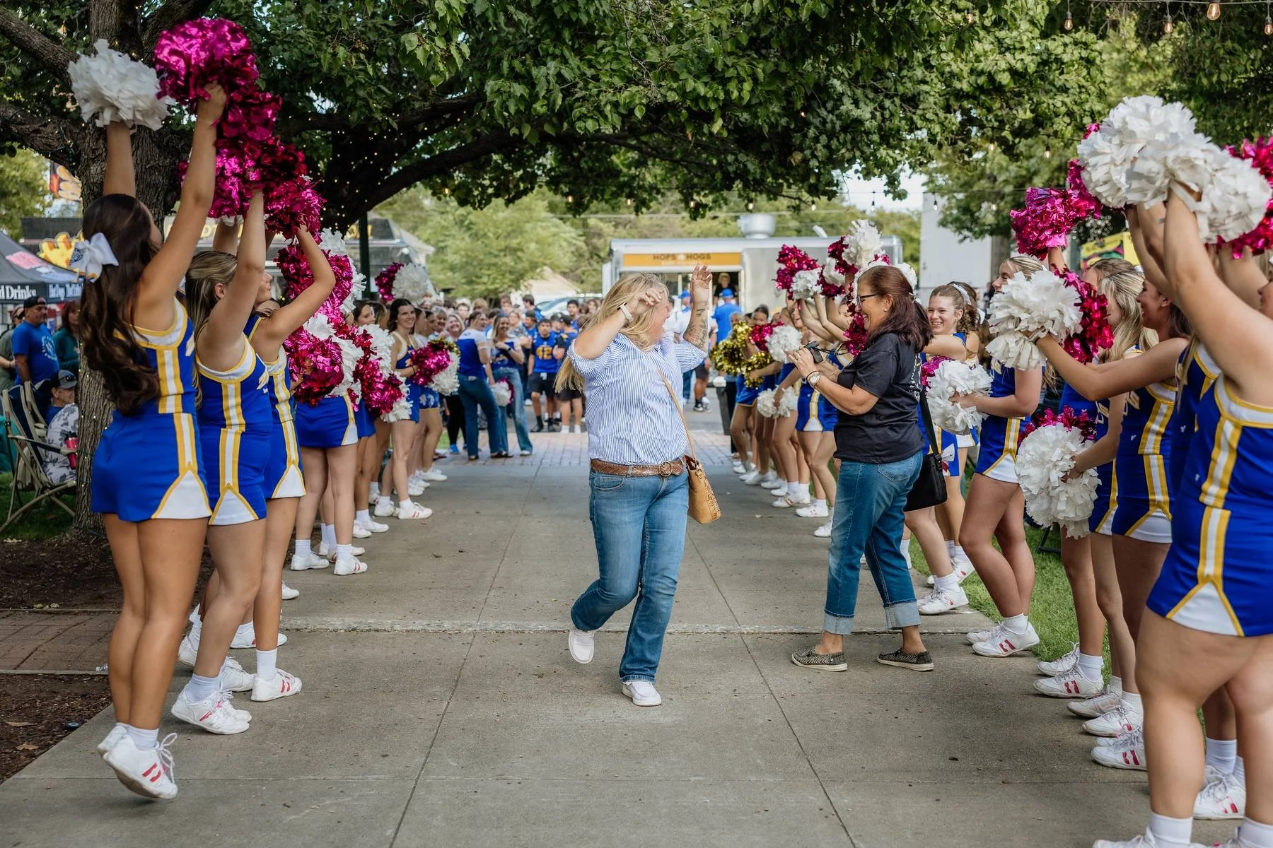 Cheerleaders forming a tunnel with pink and white pom-poms for a woman to walk through at a parade on a tree-lined street.