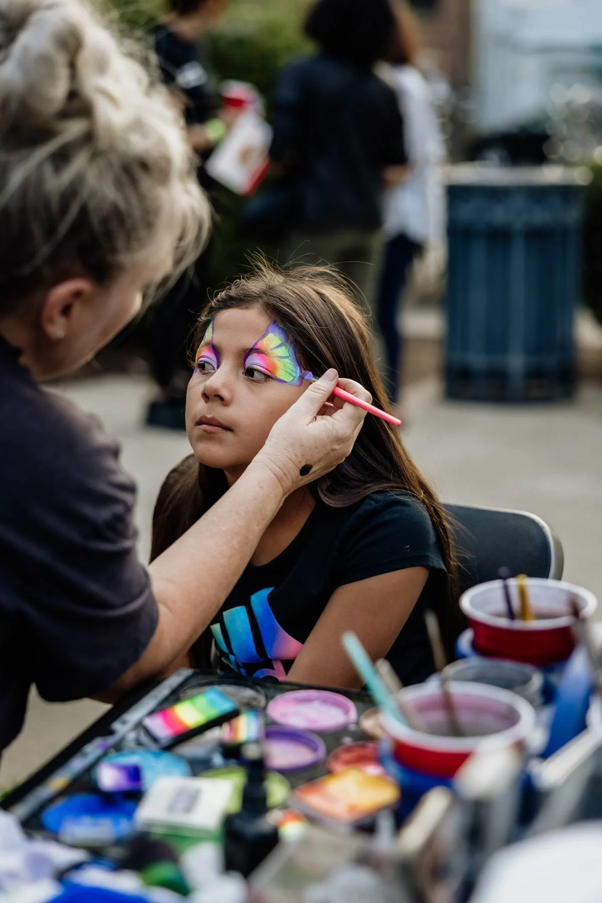 A young girl with face paint in rainbow colors is getting her face painted by an artist at an outdoor event, with face paint supplies in the foreground.