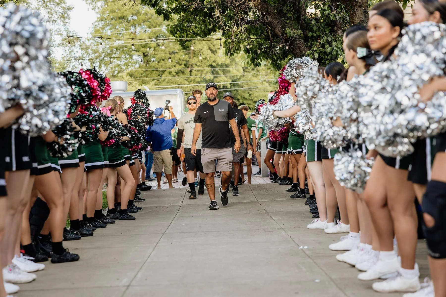 A group of cheerleaders forming a tunnel with pom poms on either side, with a man walking through in the center, during an outdoor event.
