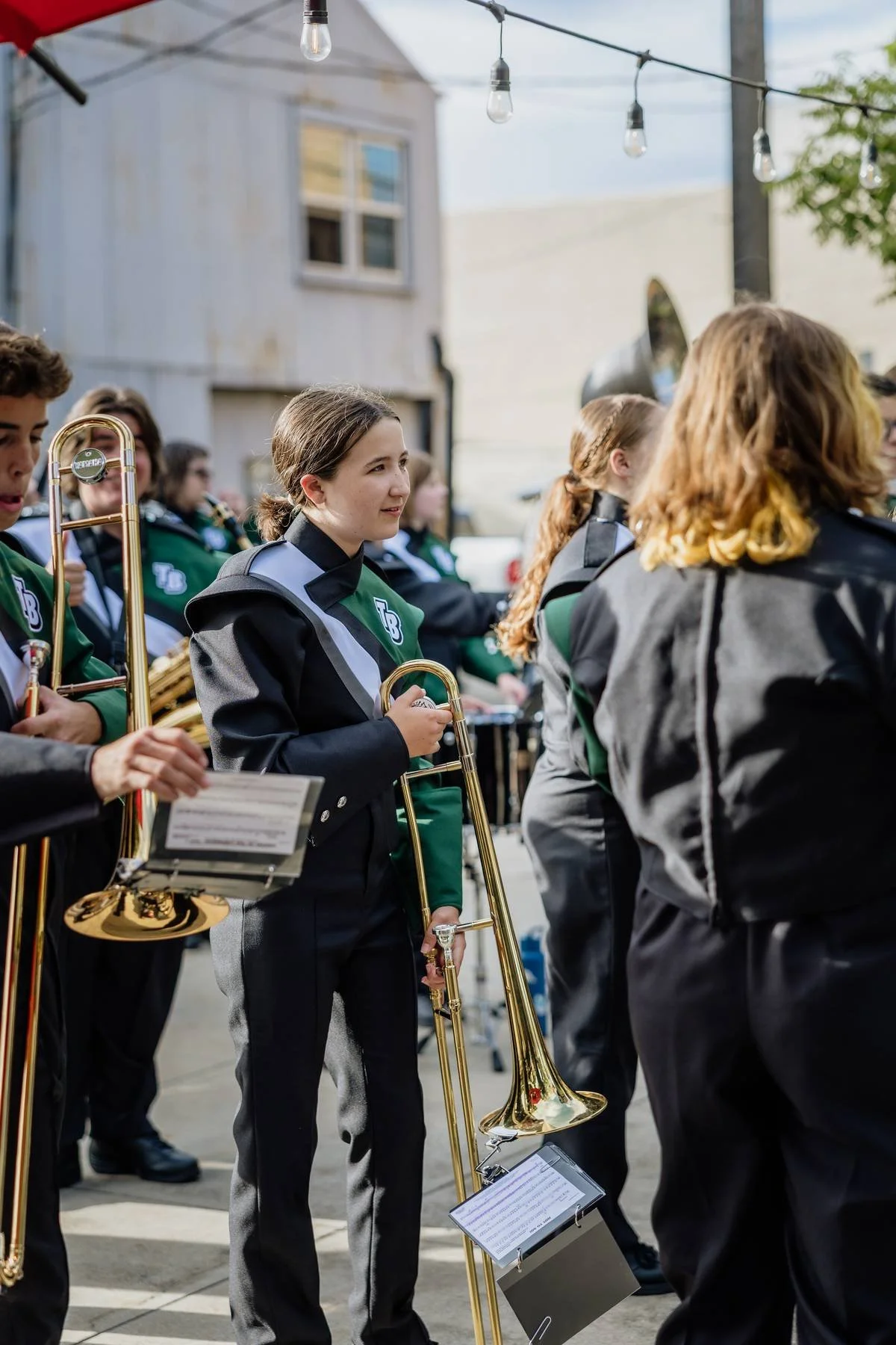 Young female musician holding a brass trombone, part of a marching band outdoors during daytime.
