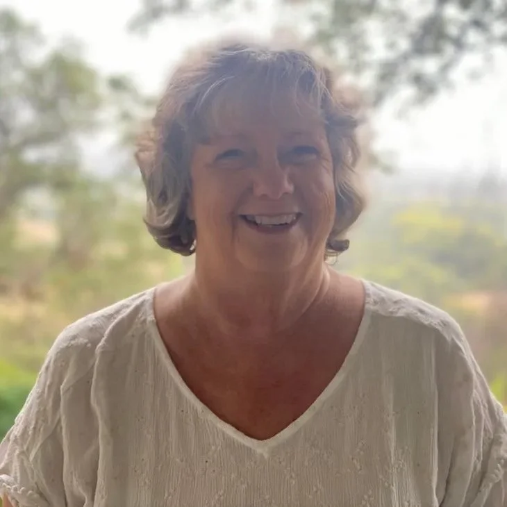 Smiling older woman with short gray hair wearing a cream-colored blouse standing outdoors with blurred trees in the background.