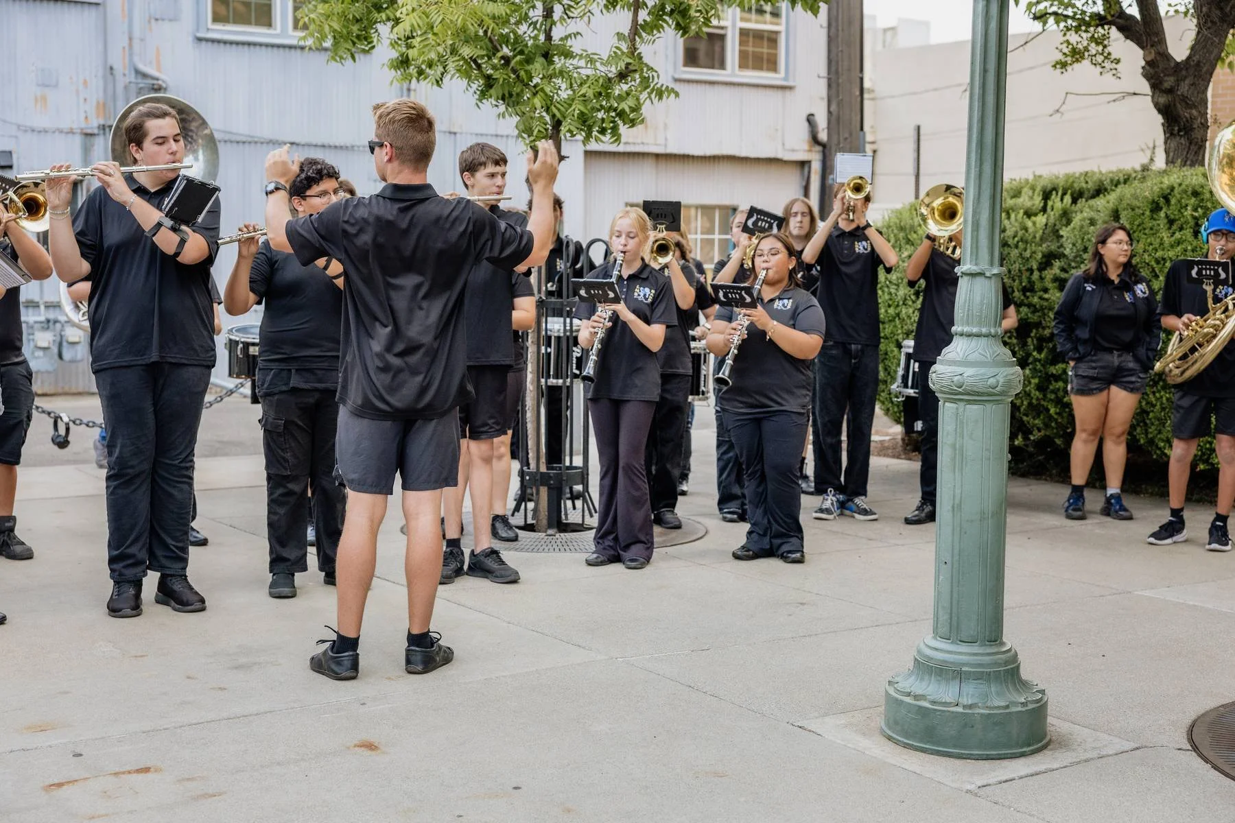 A group of young students performing in a school band outdoors, with several students playing flutes, trumpets, saxophones, and drums.