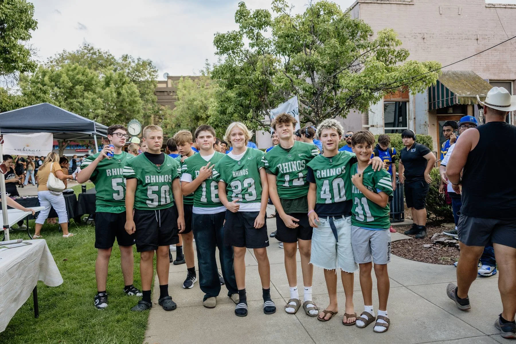 Group of young boys in green football jerseys posing together outdoors on a sidewalk, with fans and trees in the background.