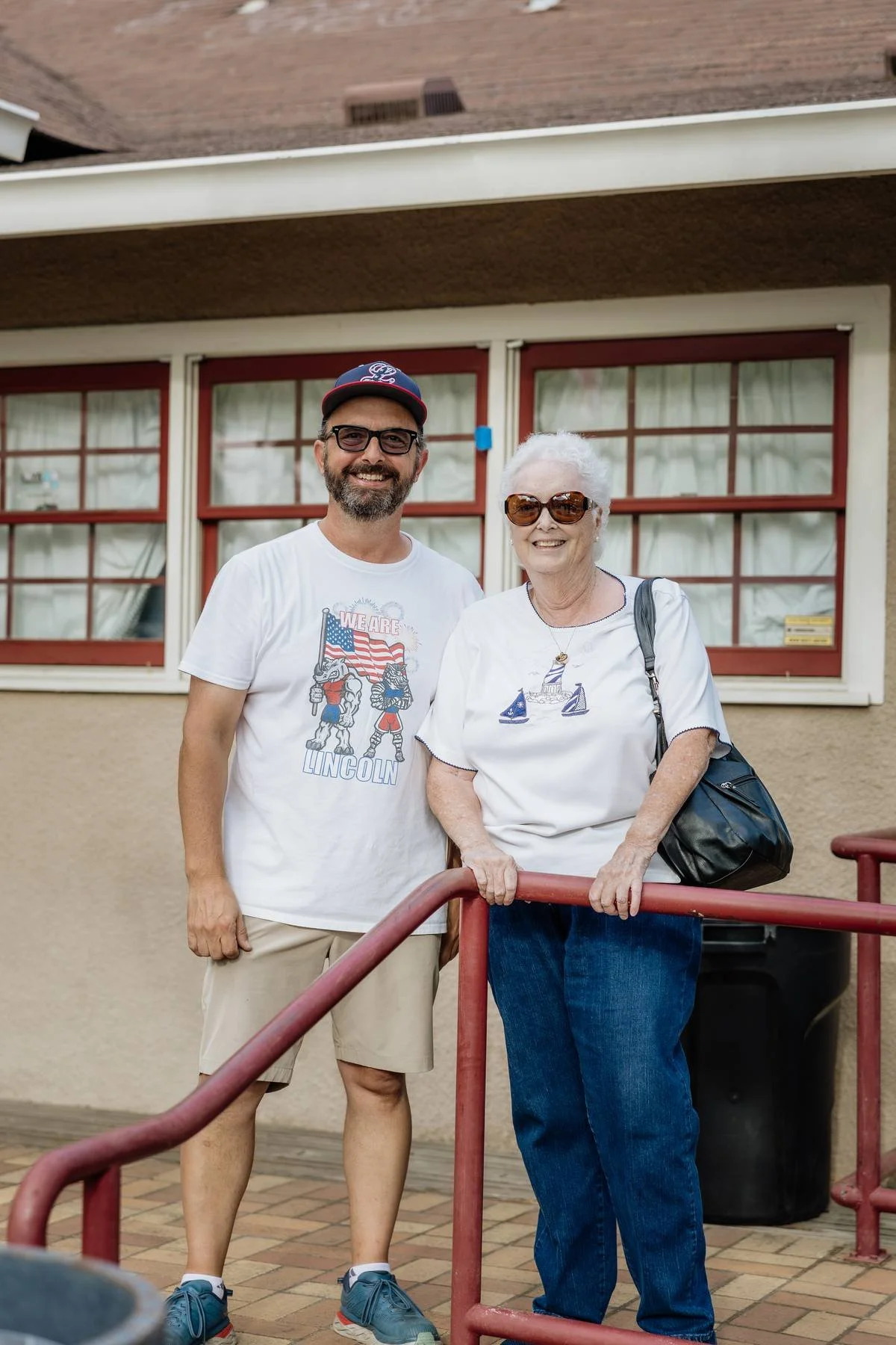 A man and an elderly woman standing together outside a building, both smiling and looking at the camera. The man is wearing a white T-shirt with a graphic and text, khaki shorts, sunglasses, and a baseball cap. The woman has white hair, large sunglas