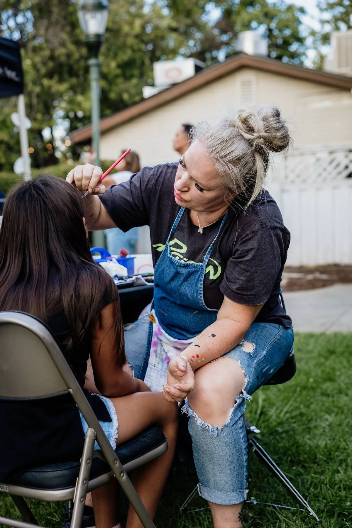 A woman with gray hair styled in a messy bun is painting a child's face outdoors. The woman is wearing a black T-shirt, a blue apron, and ripped jeans, with colorful paint on her arm. The child is sitting on a chair with long dark hair, wearing a sle