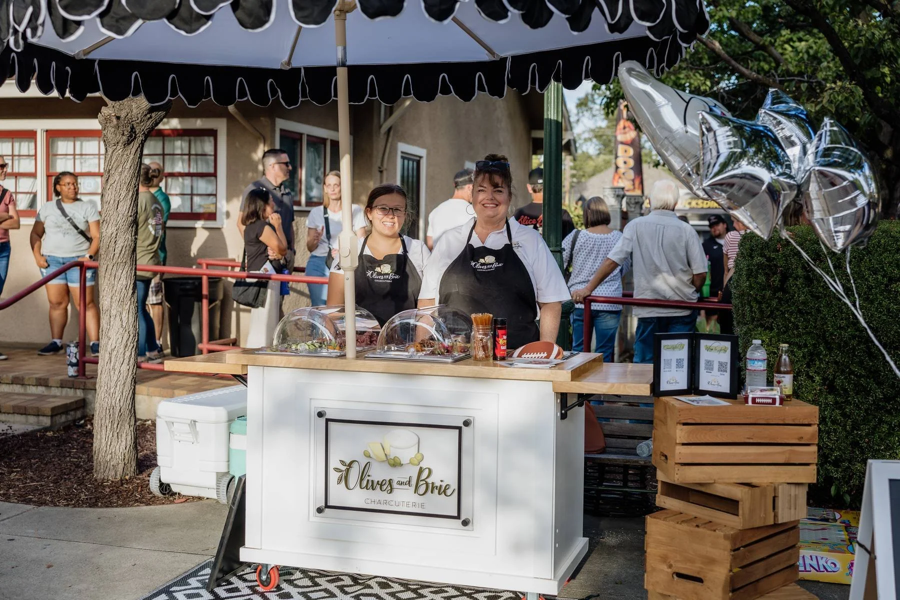 Two women standing behind a stand labeled 'Olives and Brie Charcuterie' at an outdoor event, with balloons and people in line behind them.