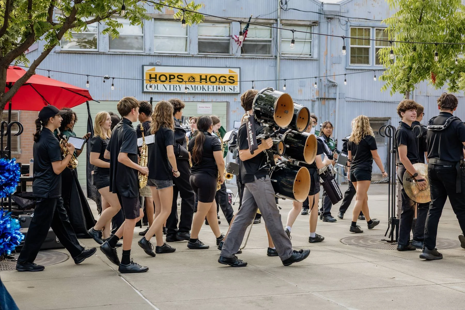 A group of young musicians walking outdoors, holding various musical instruments, with a large photograph lens on a camera operator escorting them, at a venue named 'HOPS & HOGS - COUNTRY SMOKEHOUSE.'