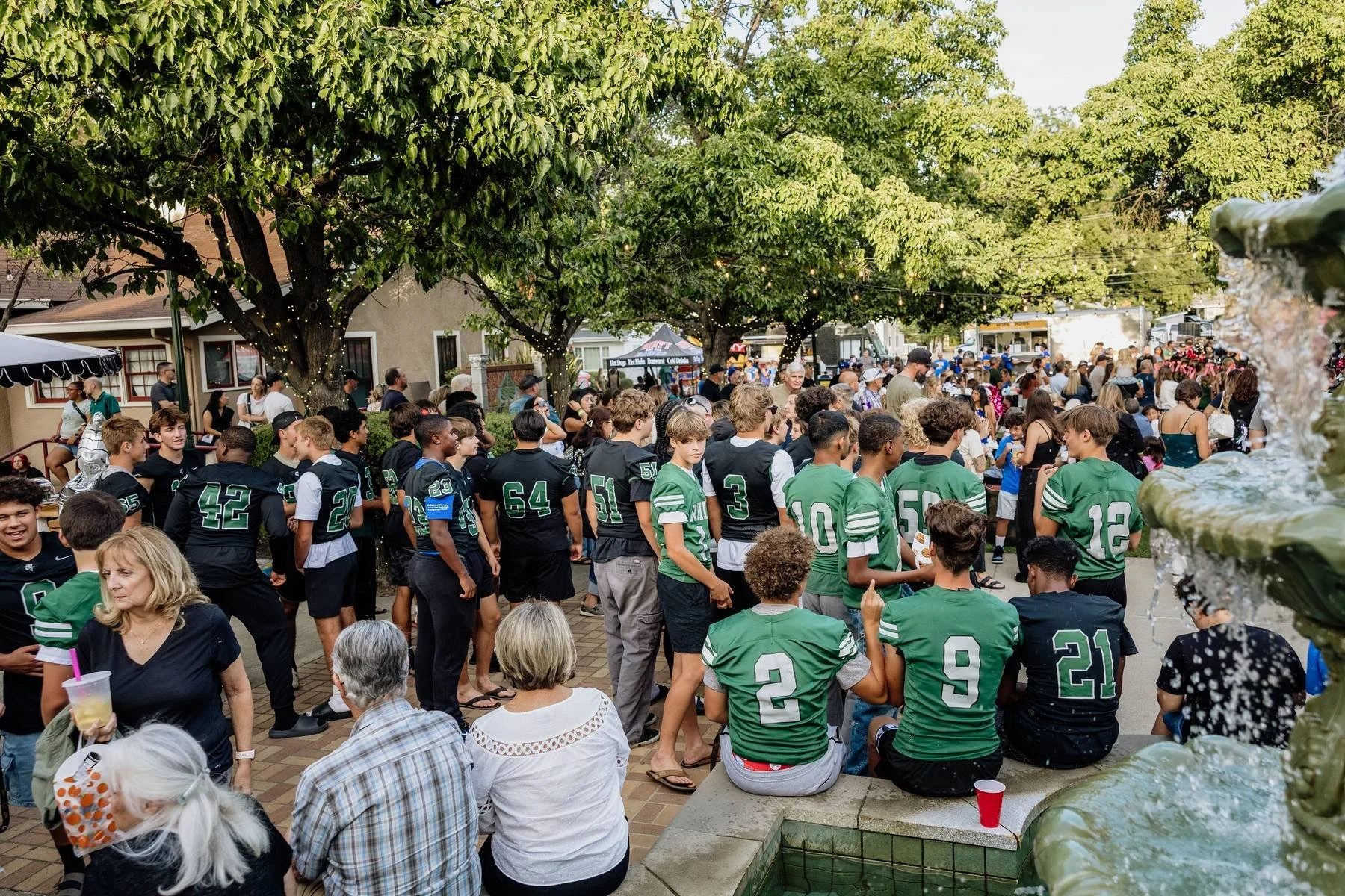 A gathering of people in a park with large trees, some connected by string lights. People are standing and sitting, some wearing football jerseys in black and green. There are food and drink vendors and a fountain in the foreground. The crowd appears